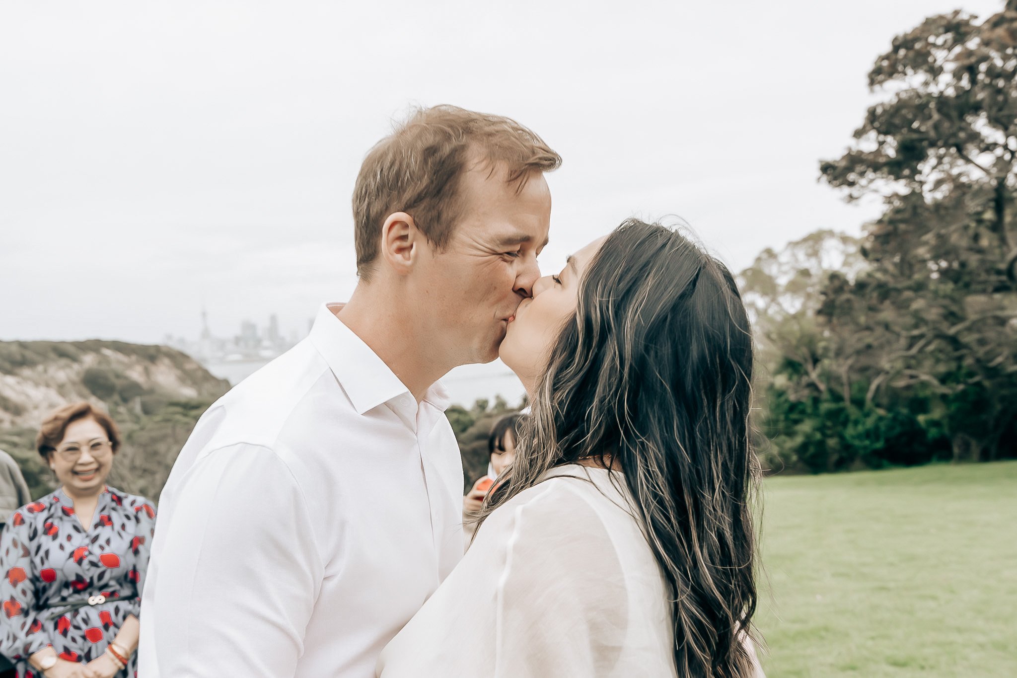 A couple is sharing a kiss outdoors, with a woman in the background smiling and wearing a patterned dress. The scene appears to be at a park or garden on a cloudy day.