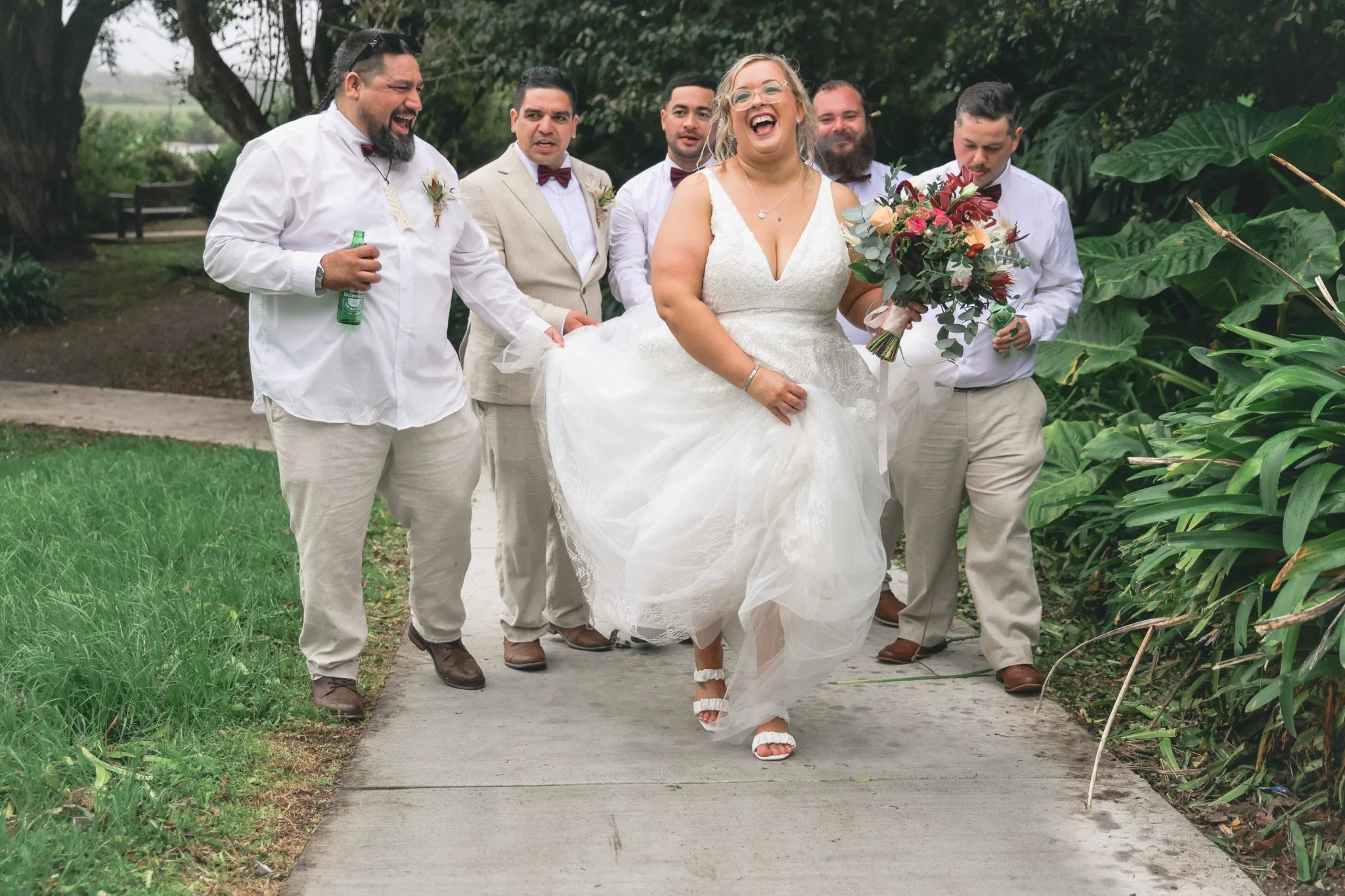 A bride in a white wedding dress holding a bouquet, walking with a group of groomsmen dressed in white shirts, beige pants, and maroon bow ties outdoors on a sidewalk surrounded by greenery.