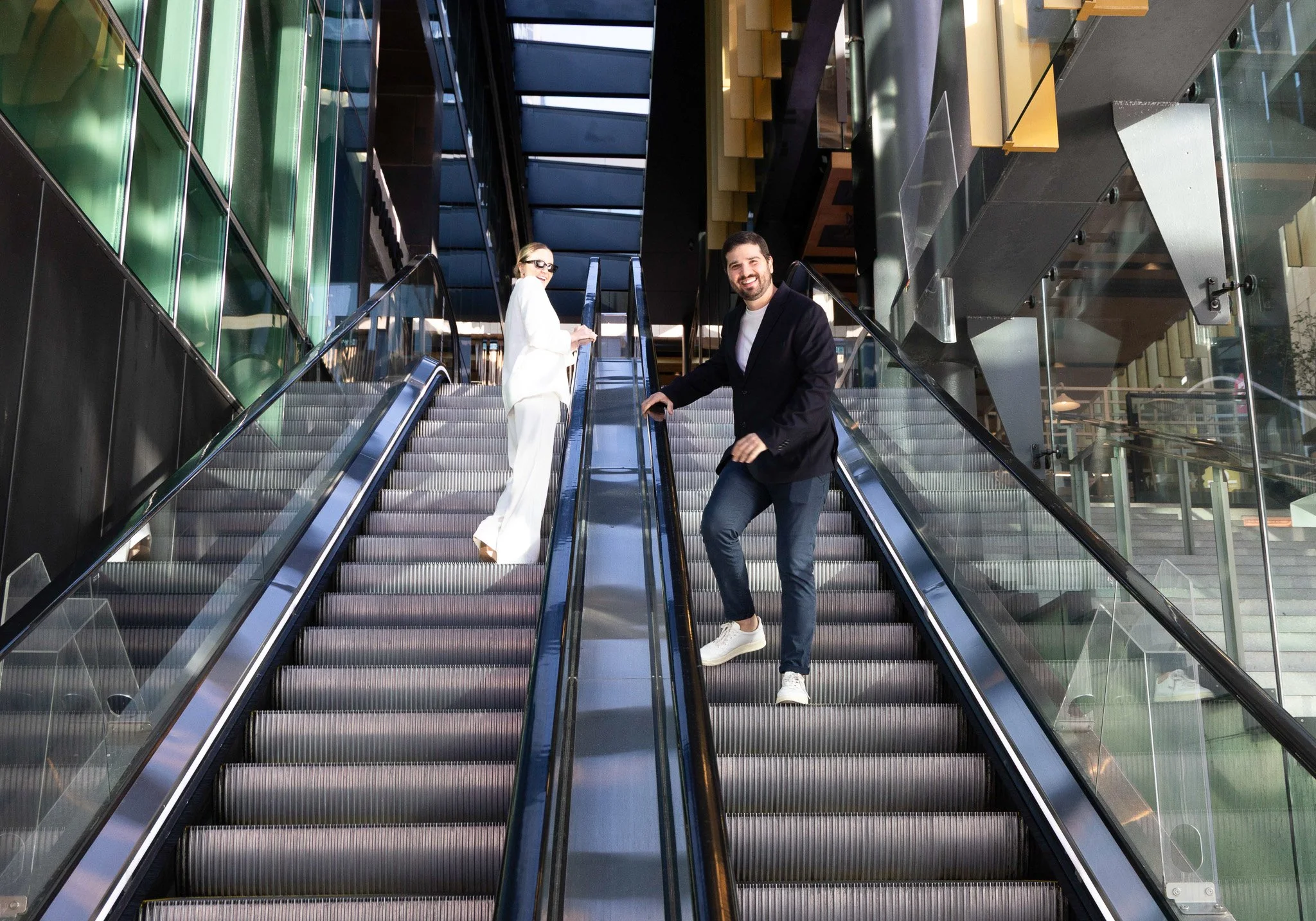 A man and woman riding escalators in a modern building, with glass walls and a staircase visible in the background, all illuminated by natural light from ceiling windows.