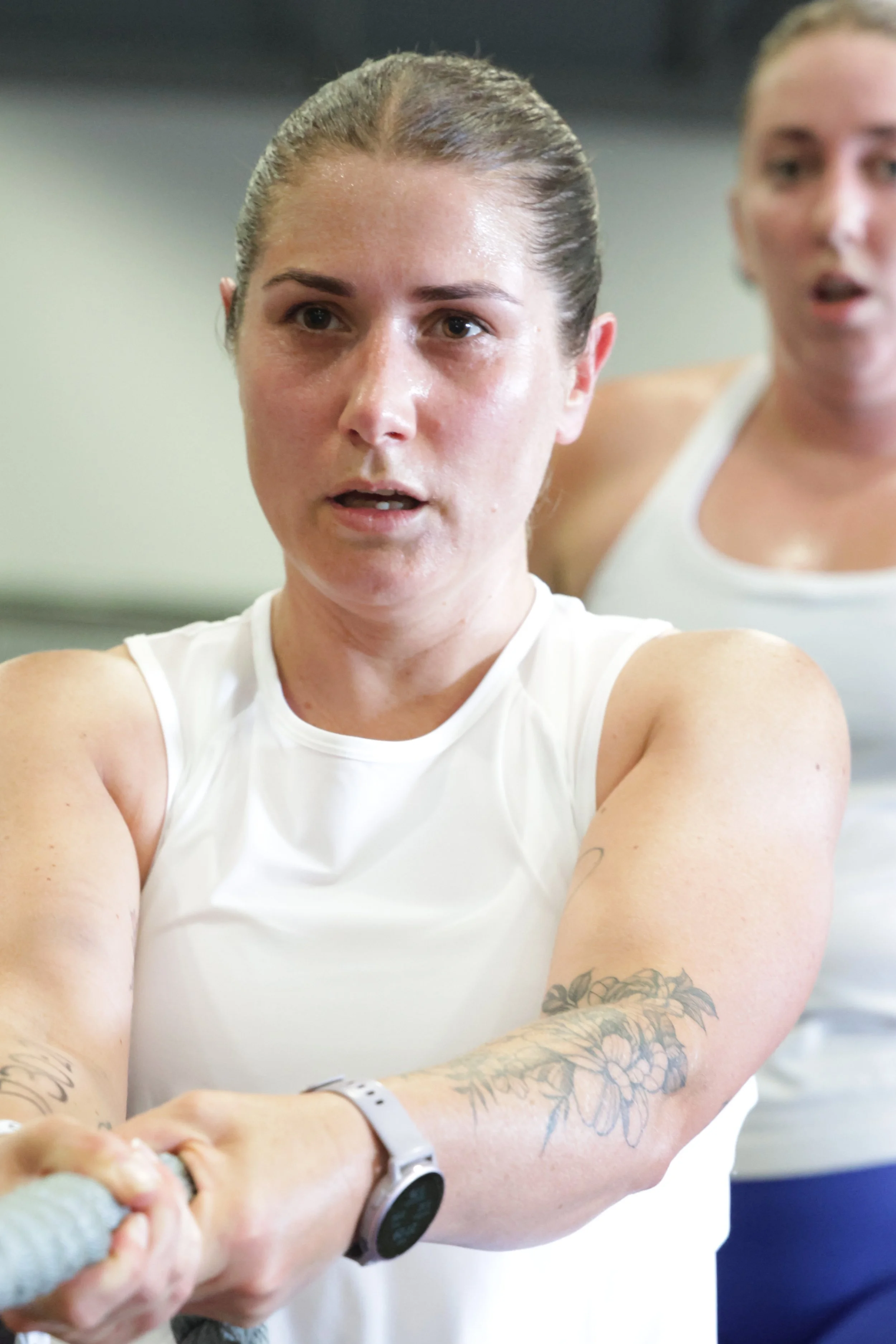 A woman with brown hair tied back, wearing a white tank top and a smartwatch, is gripping a rope during a workout, with tattoos on her arms. Another woman is slightly blurred in the background, also in a white tank top.