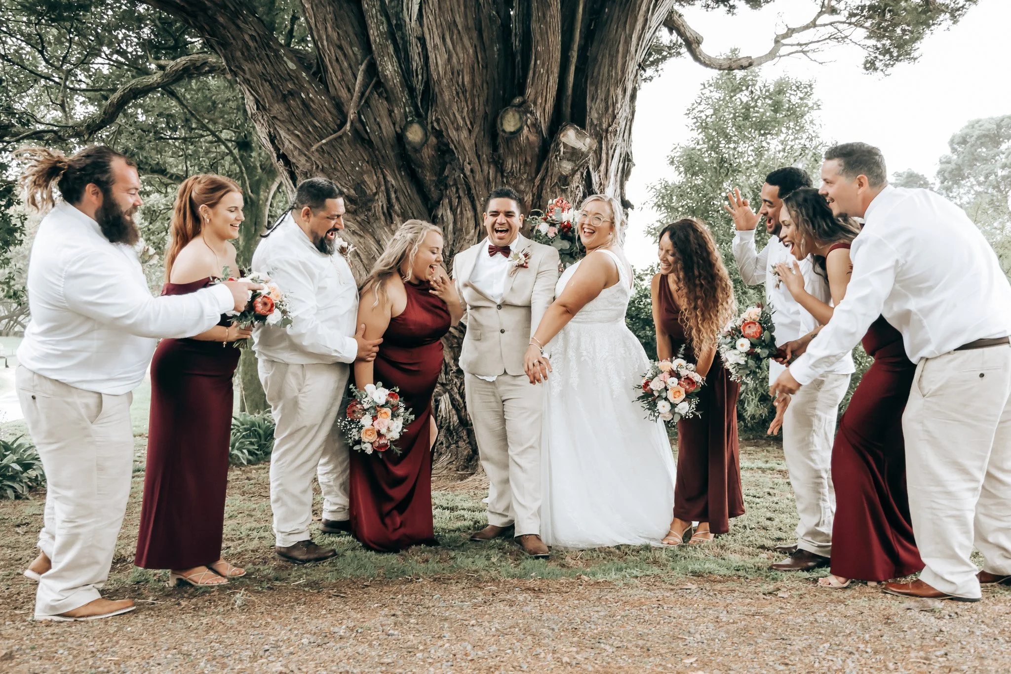 A group of people in wedding attire is gathered outdoors, laughing and celebrating under a large tree.