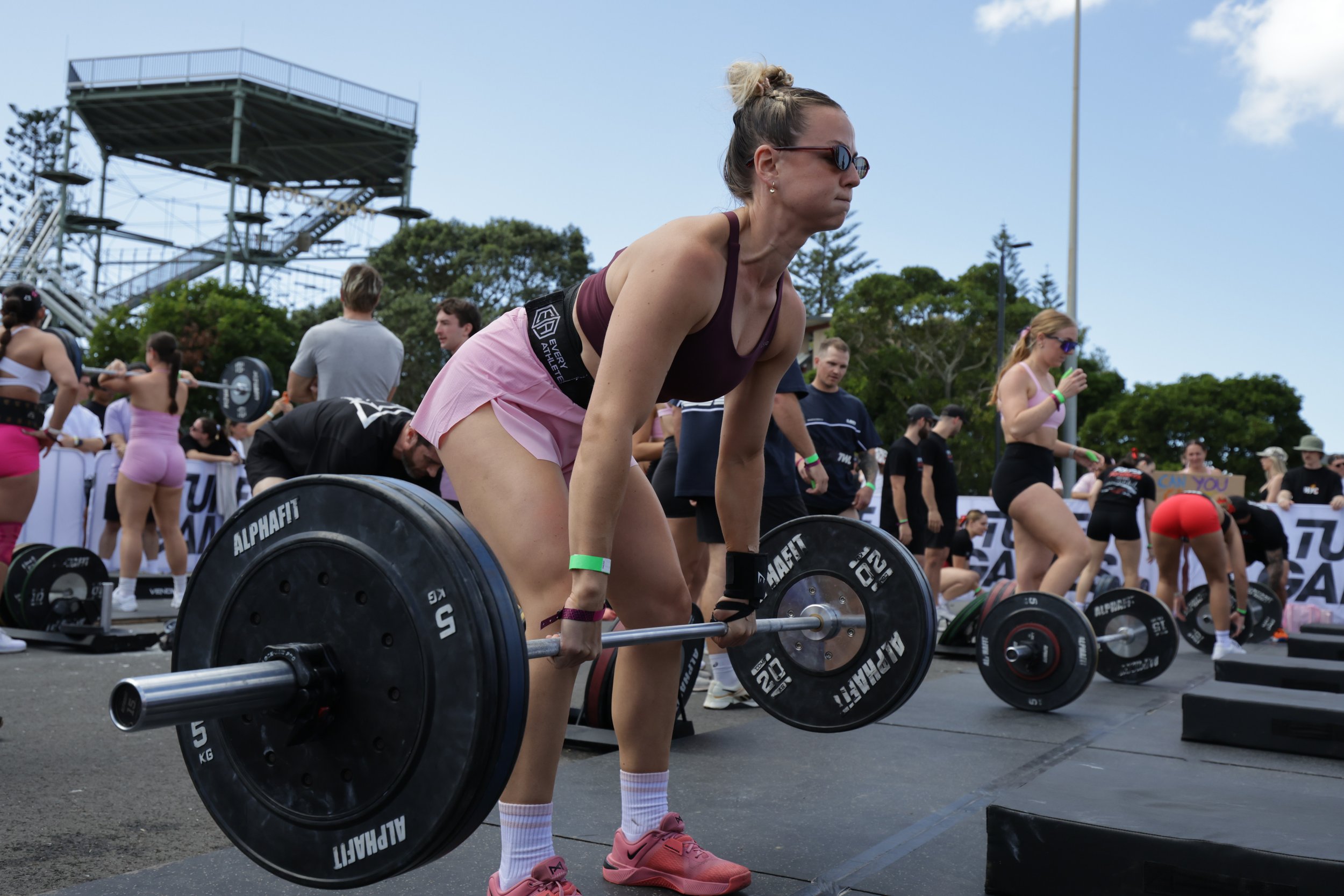 Woman wearing sunglasses and pink shoes lifting a barbell during an outdoor weightlifting event with spectators in the background.