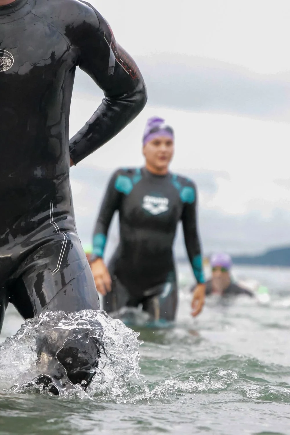 People in wetsuits participating in a swimming event in the water, with a cloudy sky in the background.