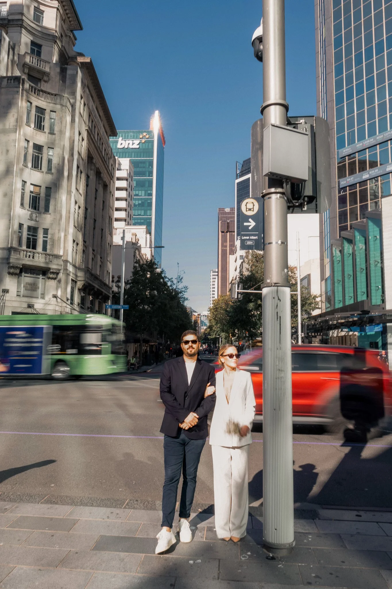 Two people walking on city sidewalk crossing a busy street with tall buildings, including a BNZ bank building, in the background. One man and one woman, dressed up, are holding arms.