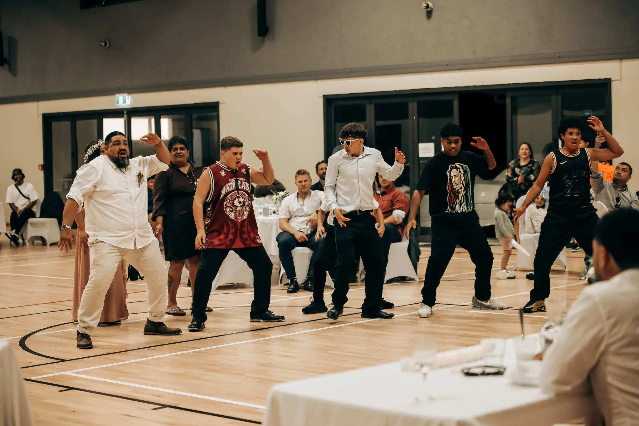 Group of young people dancing in a gymnasium at a gathering or party, with tables and guests in the background.