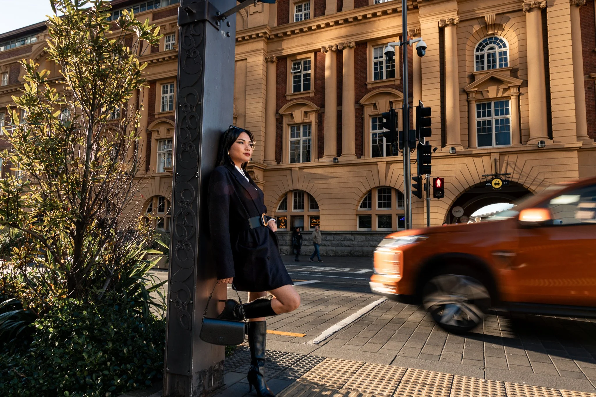 A woman in a black blazer with rolled sleeves, wearing black knee-high boots, sits on a sidewalk curb next to a decorative street lamp pole and a small green tree in an urban area. Behind her, there's a large, ornate multi-story building with arched 