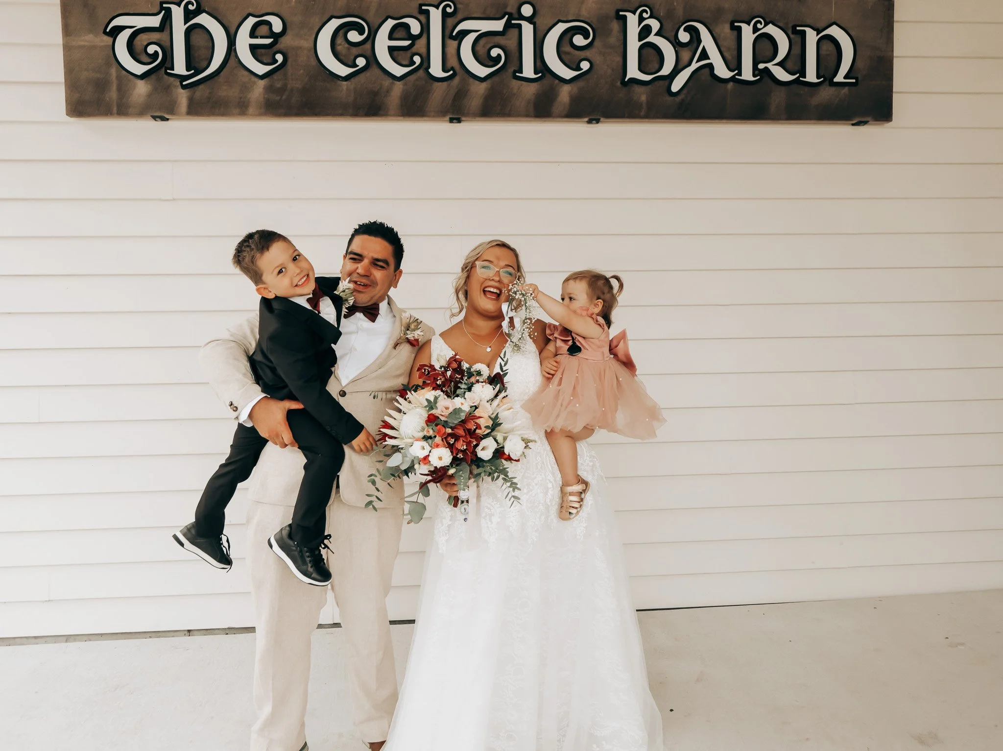 A smiling bride in a white lace wedding dress holding a bouquet, standing next to a man in a beige suit with a boutonniere, and two children dressed in formal attire, before a white shiplap wall with a sign that reads 'The Celtic Barn' in black decor