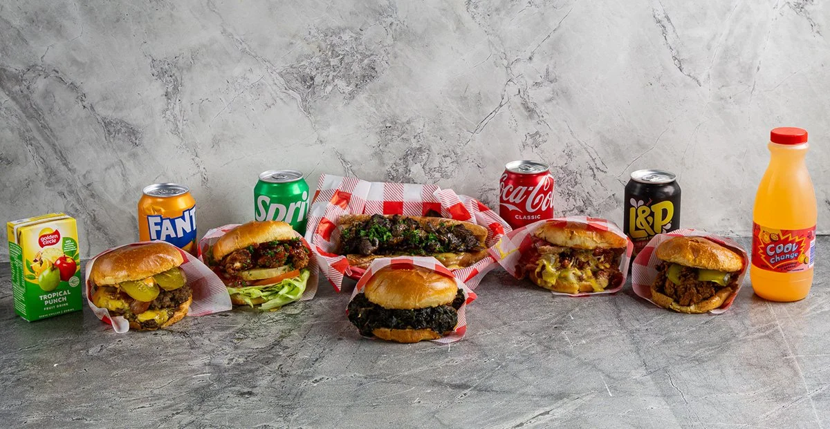 A variety of fast food items including burgers, soft drinks, juice, and a small carton of fruit juice on a gray surface with a textured gray background.