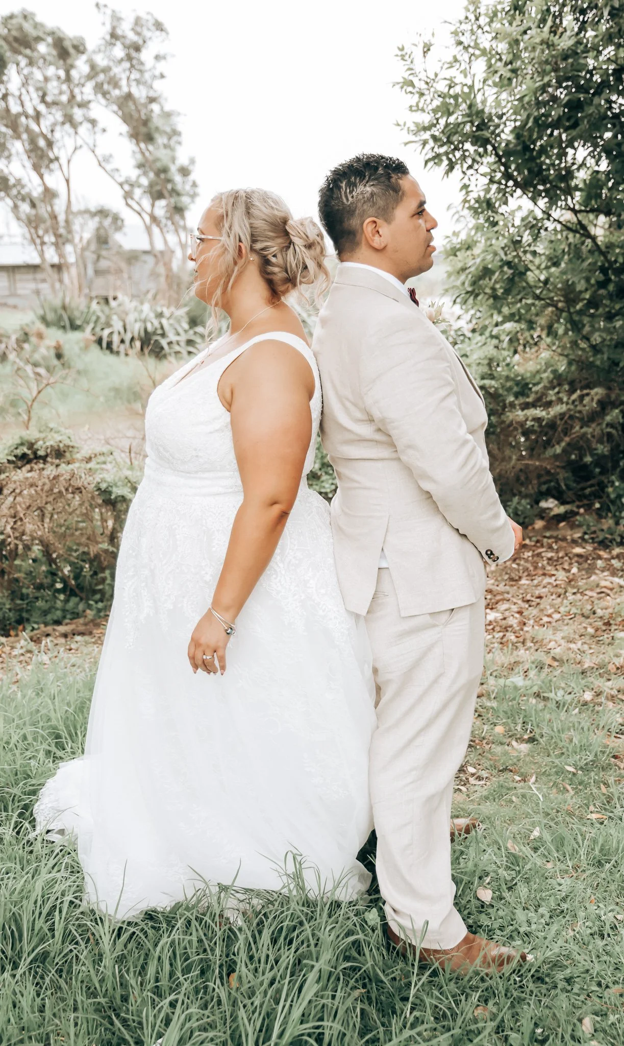 A bride and groom standing back to back outdoors during a wedding ceremony, with the bride in a white lace wedding dress and the groom in a light-colored suit, both facing away from each other amidst green trees and grass.