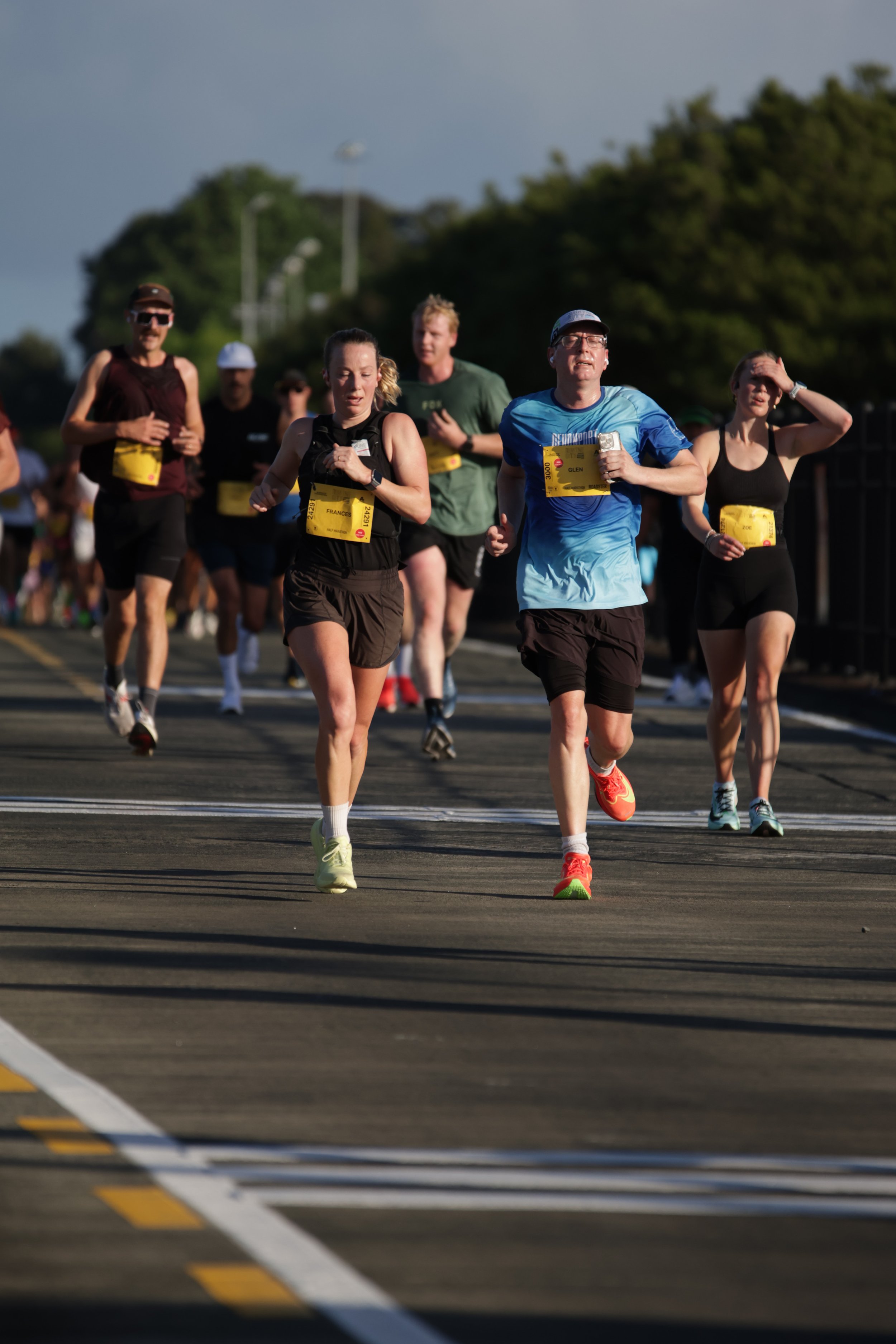 Group of runners competing in a marathon, running on a city street during daylight, surrounded by trees in the background.