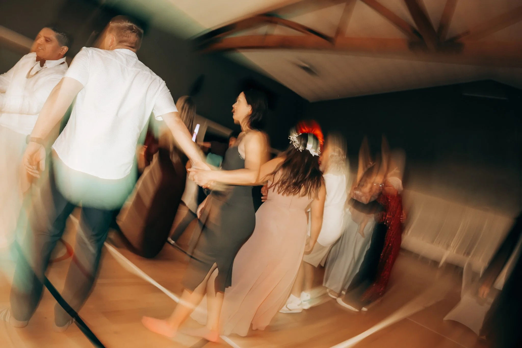 People dancing at a social gathering or party, holding hands in a conga line, with blurred motion effects in the background.