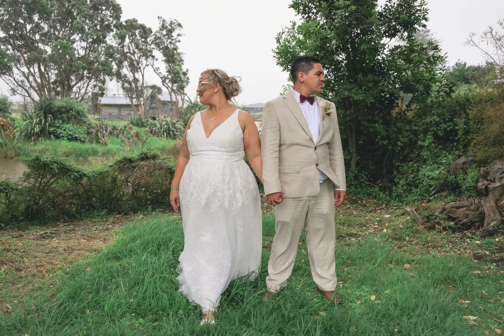 A bride and groom stand hand-in-hand outdoors, facing away from each other, in a lush green garden with trees and bushes, on a cloudy day.
