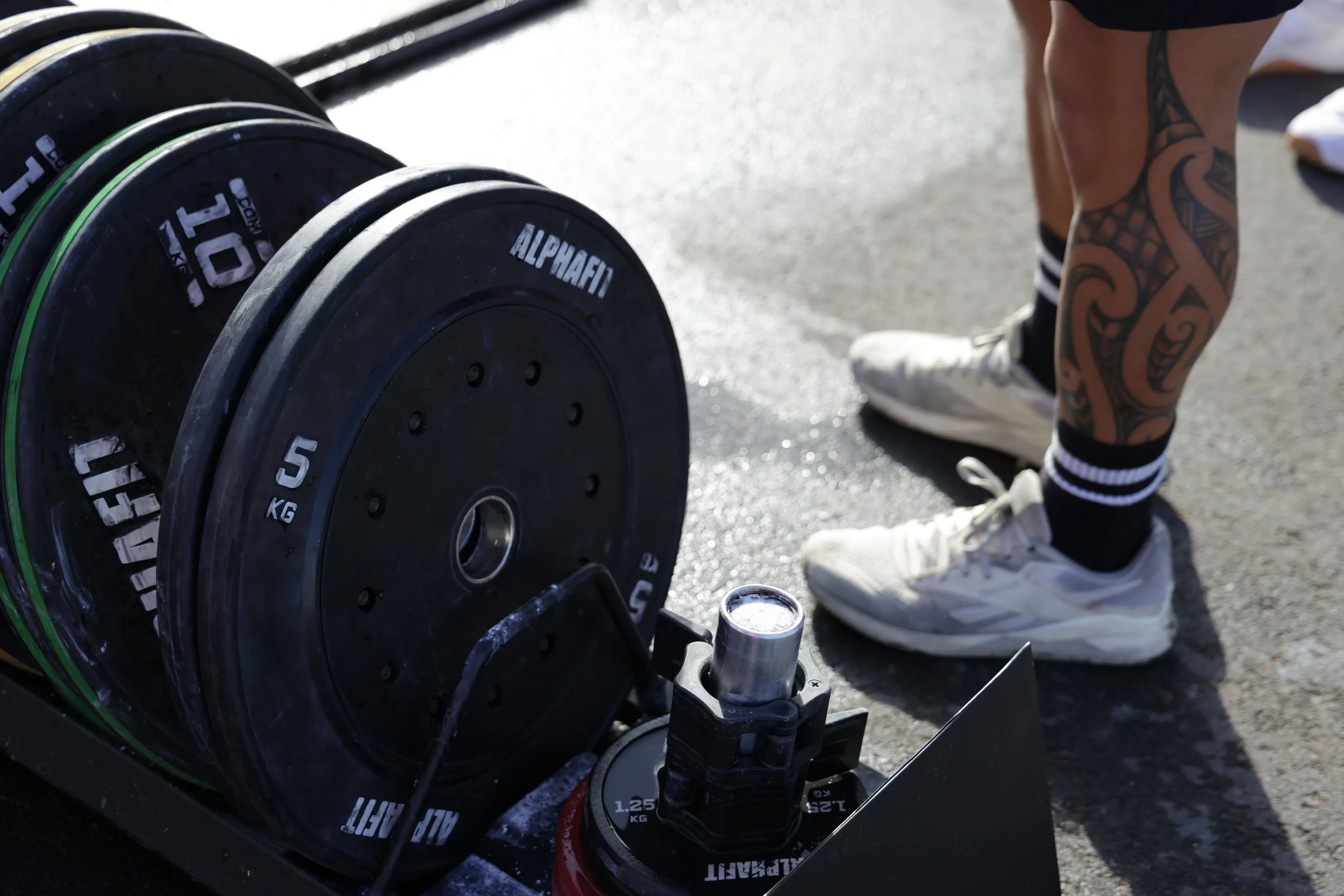 Close-up of weightlifting equipment, including black weight plates labeled 'ALPHAFIT' and '10 kg,' on a barbell. There is a person with tattoos on their left calf, wearing black shorts, black and white striped socks, and white shoes, standing nearby 
