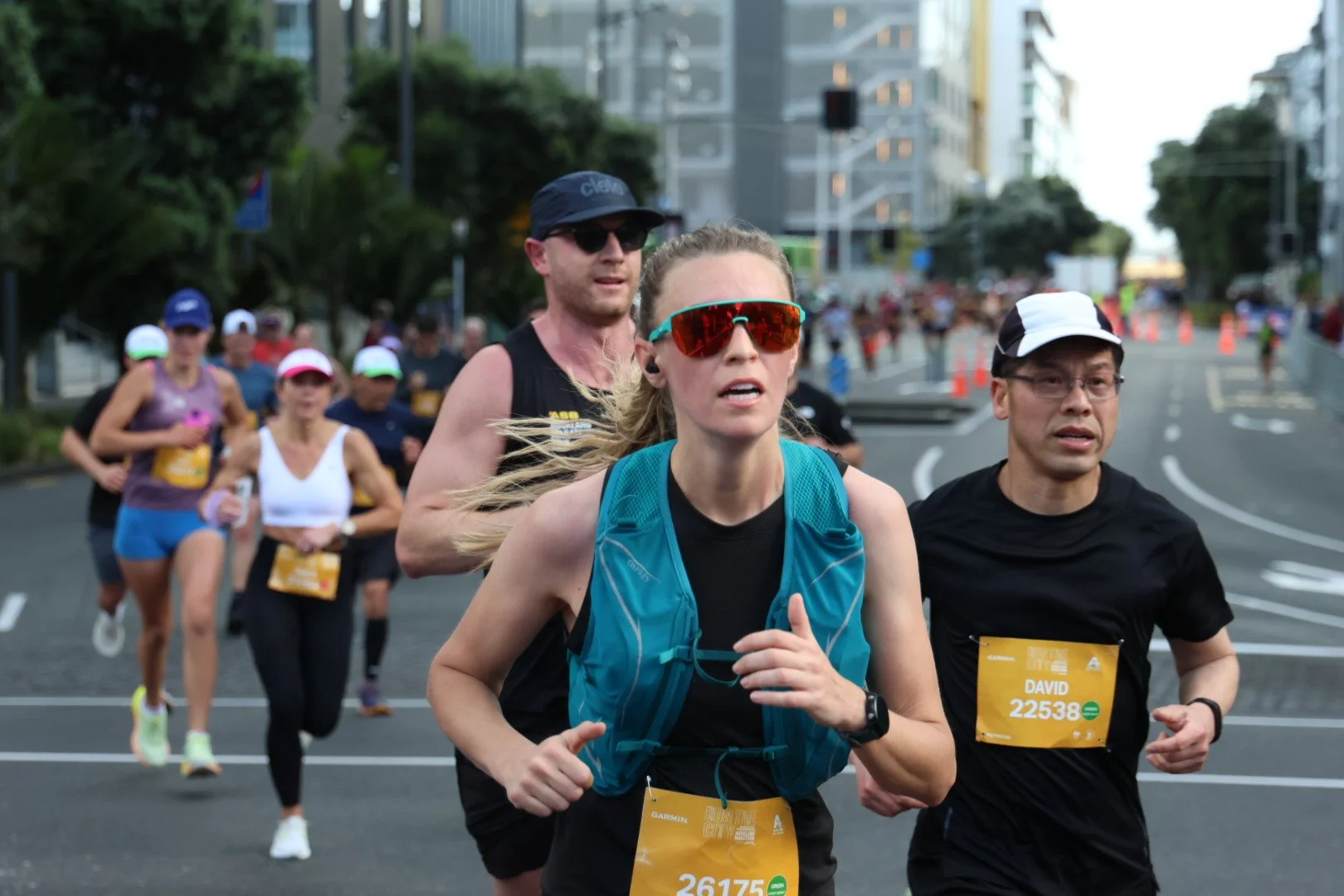 Group of runners participating in a marathon on city streets, with tall buildings, trees, and orange traffic cones in the background.