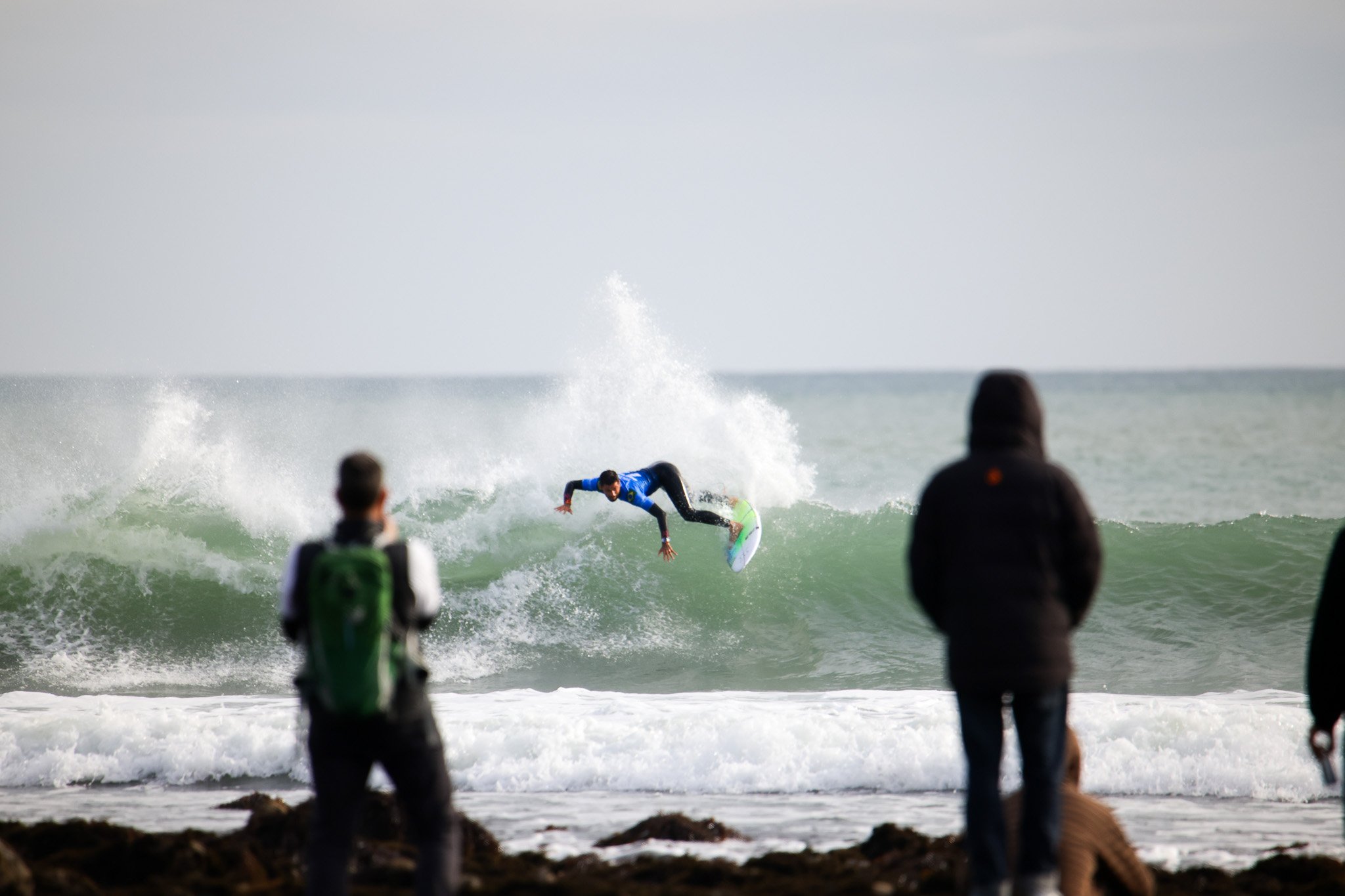A surfer riding a wave at the beach with onlookers watching from the shoreline.