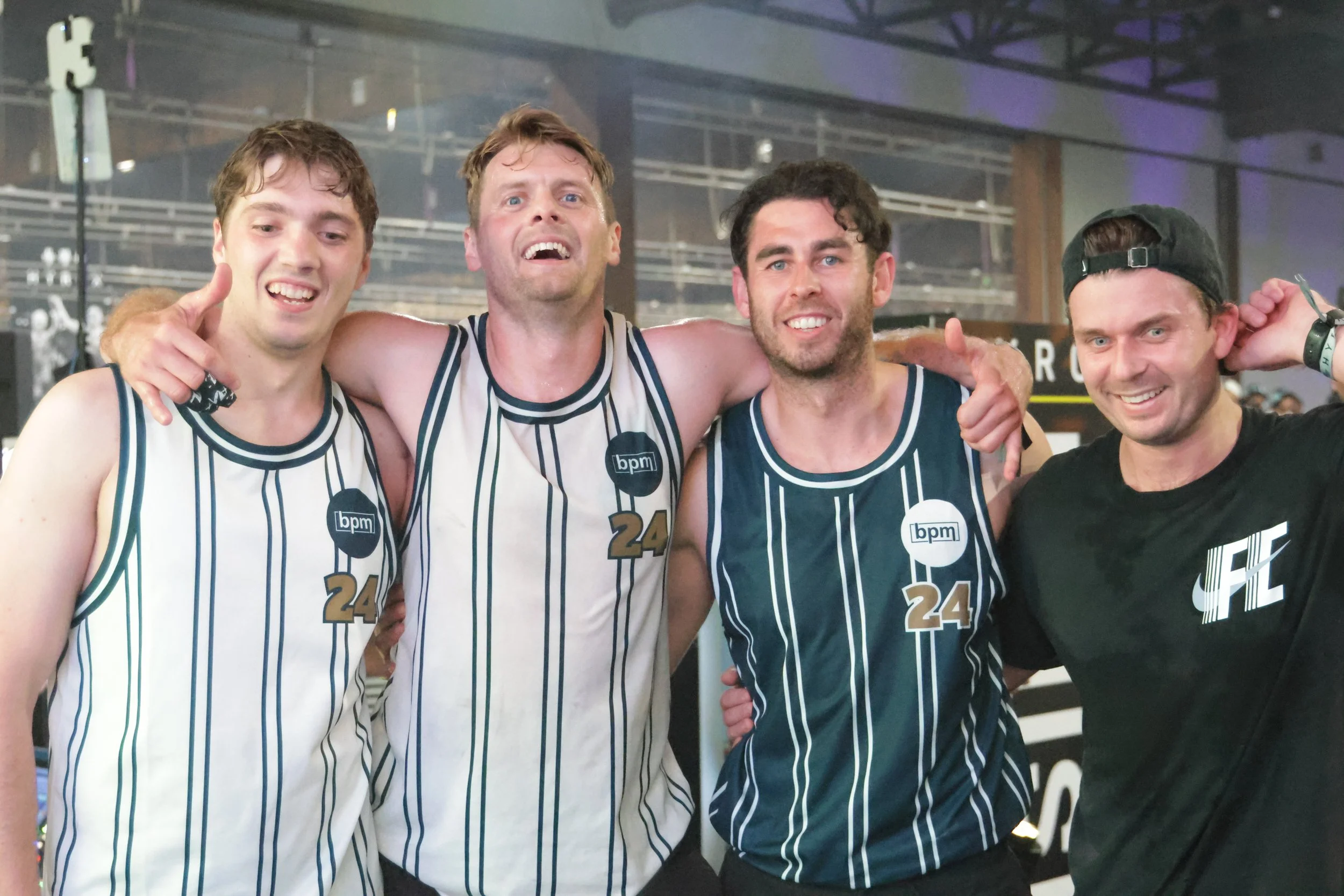 Four young men celebrating, wearing basketball jerseys with vertical stripes, posing with their arms around each other, smiling at the camera in an indoor sports arena.