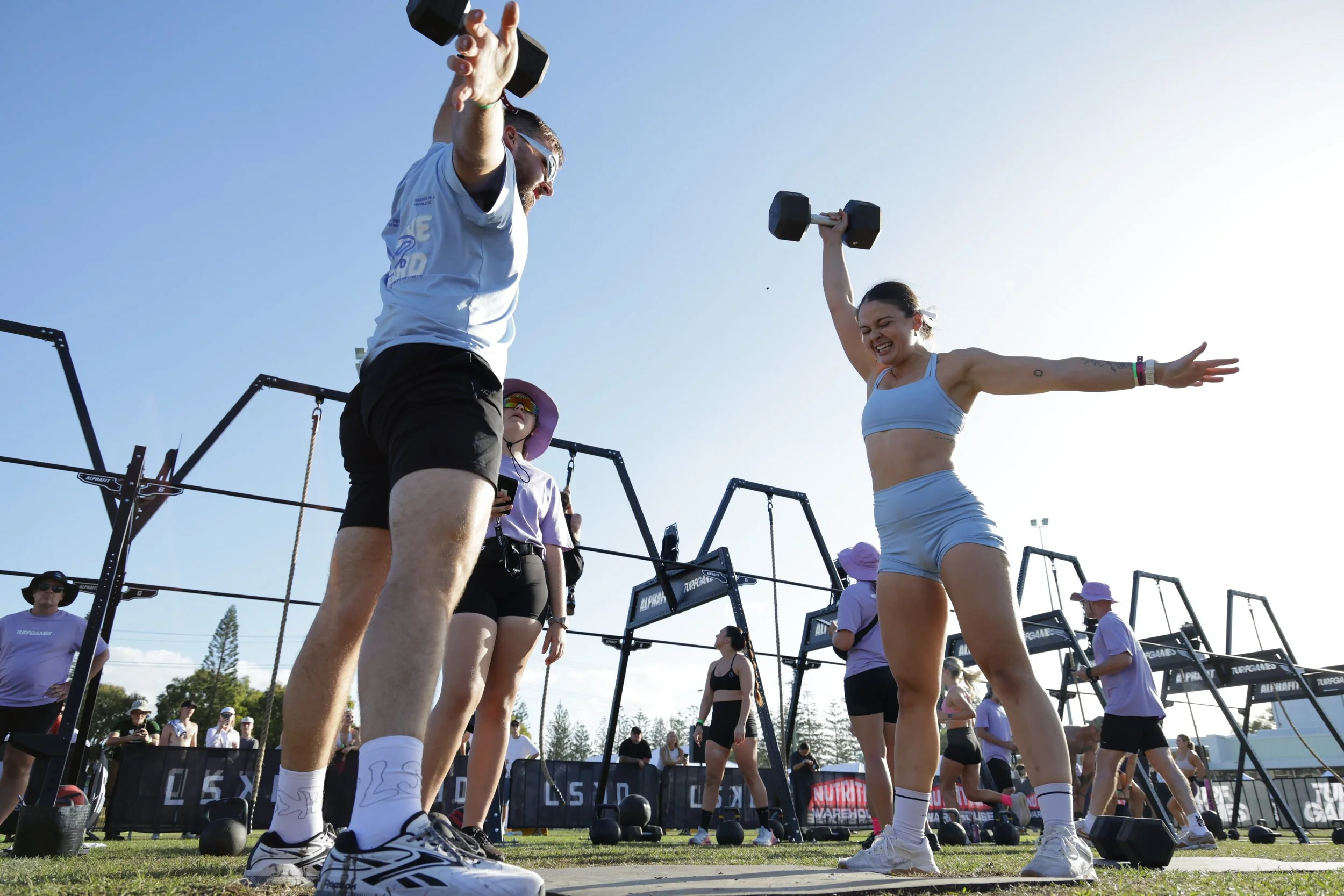 A group of people, mostly women, participating in a fitness event outdoors. They are lifting dumbbells with smiling, energetic expressions. The sky is clear and sunny, with some onlookers and event staff in the background.