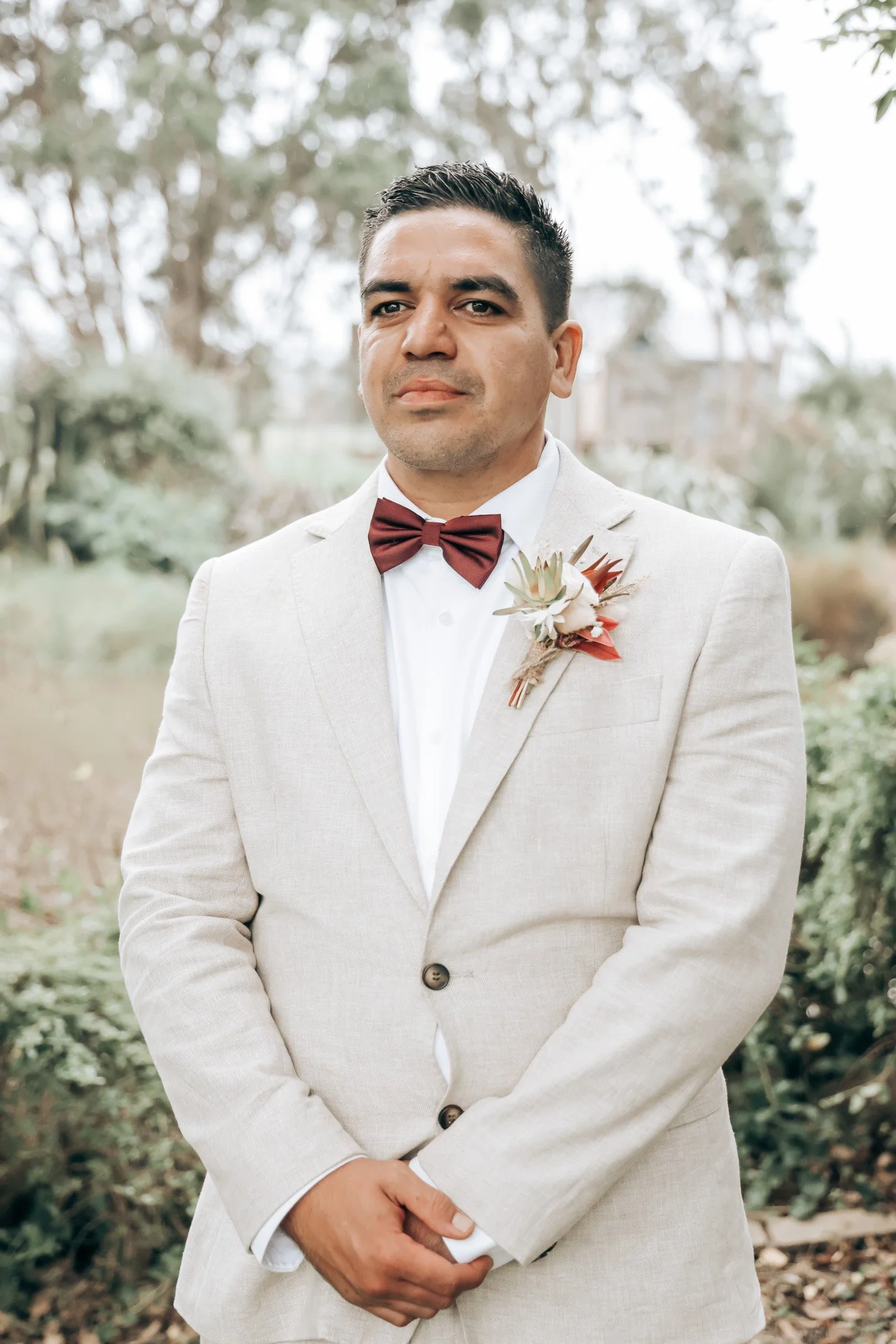 A man dressed in a light-colored suit with a maroon bow tie and boutonniere, standing outdoors with trees and blurred background.