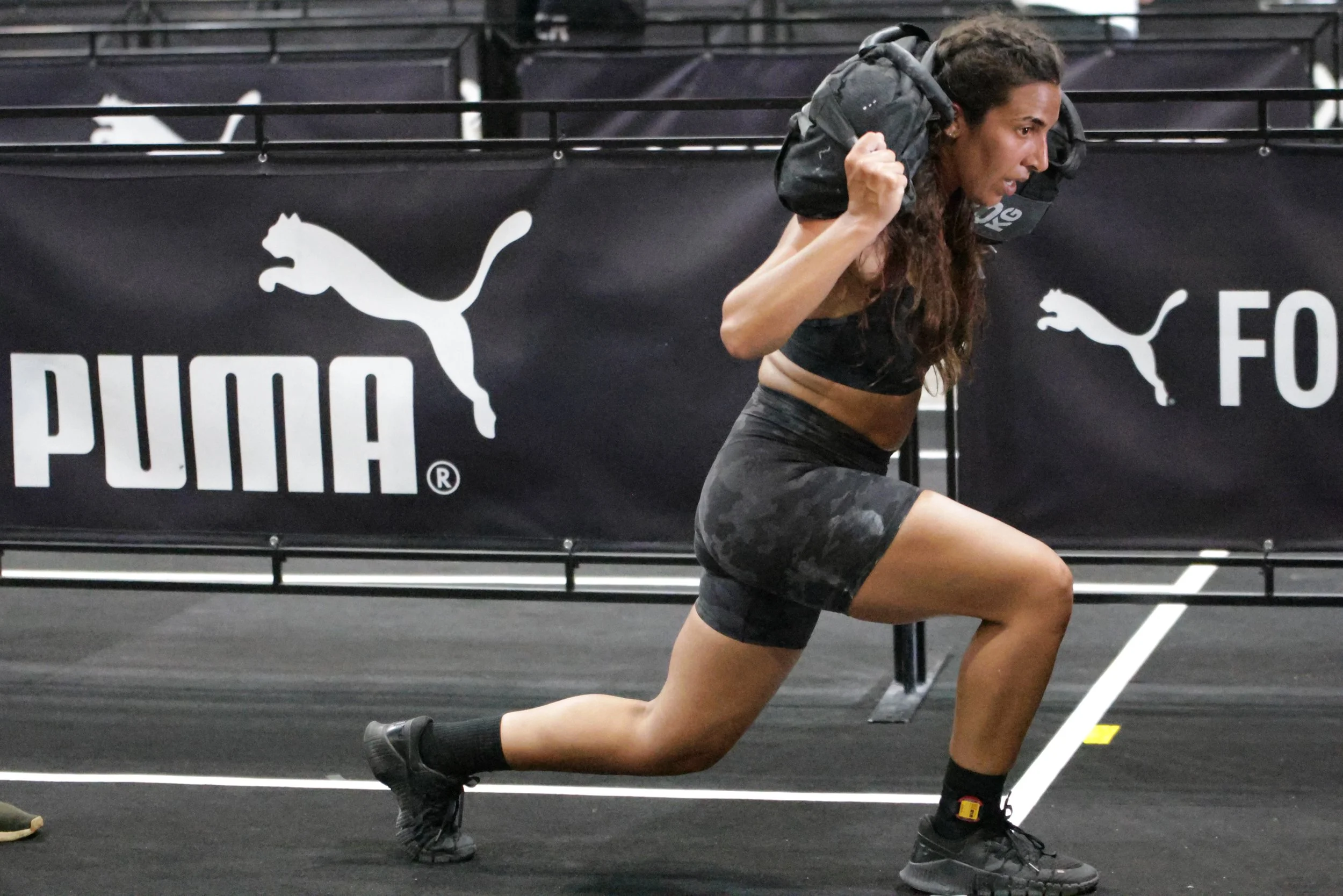 A woman doing a lunge exercise at a gym, holding a backpack on her shoulders, with black athletic wear and the Puma logo visible in the background.