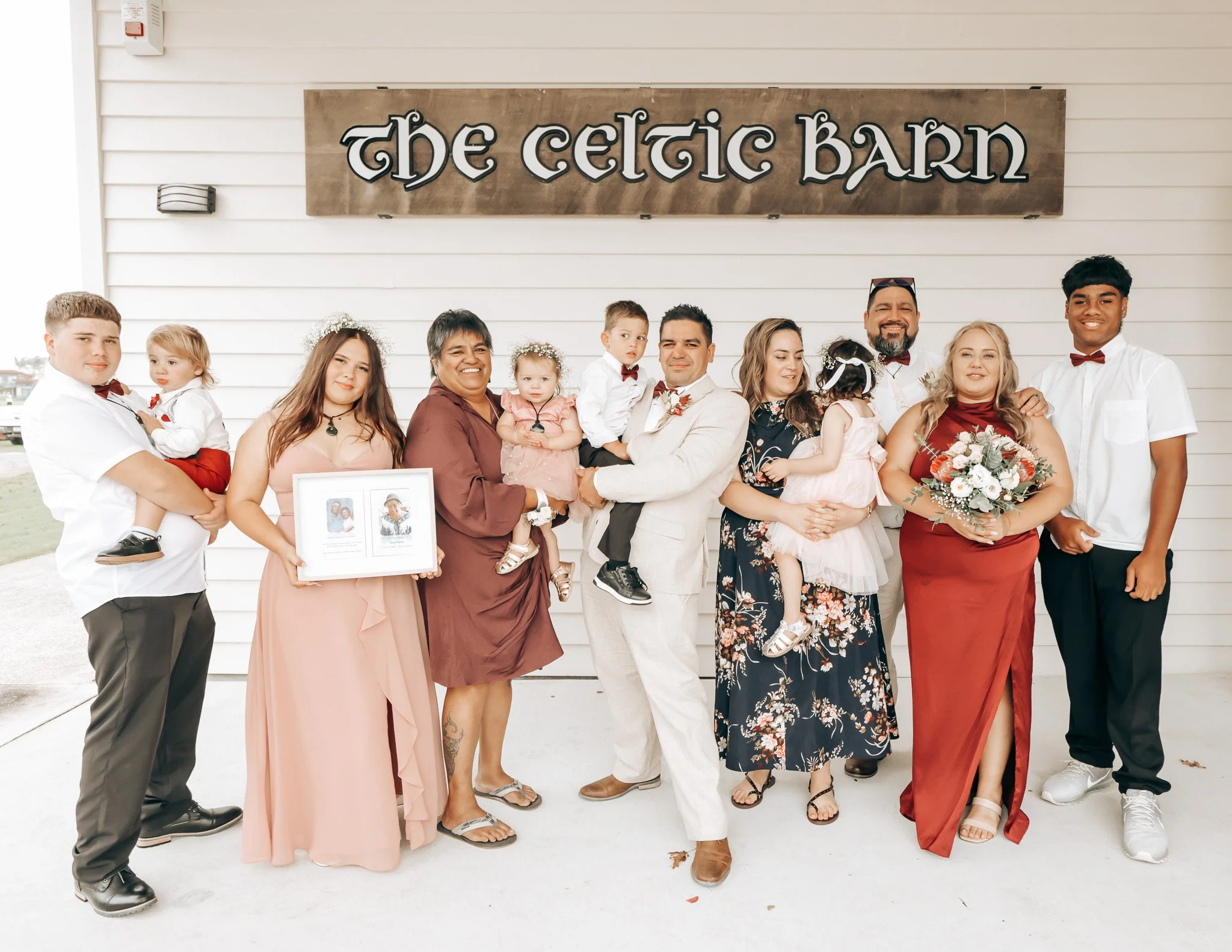A group of people, including children and adults, standing together in front of a sign that reads 'The Celtic Barn'. They are dressed in semi-formal attire, with some women holding bouquets, and some people wearing flower crowns. The background featu
