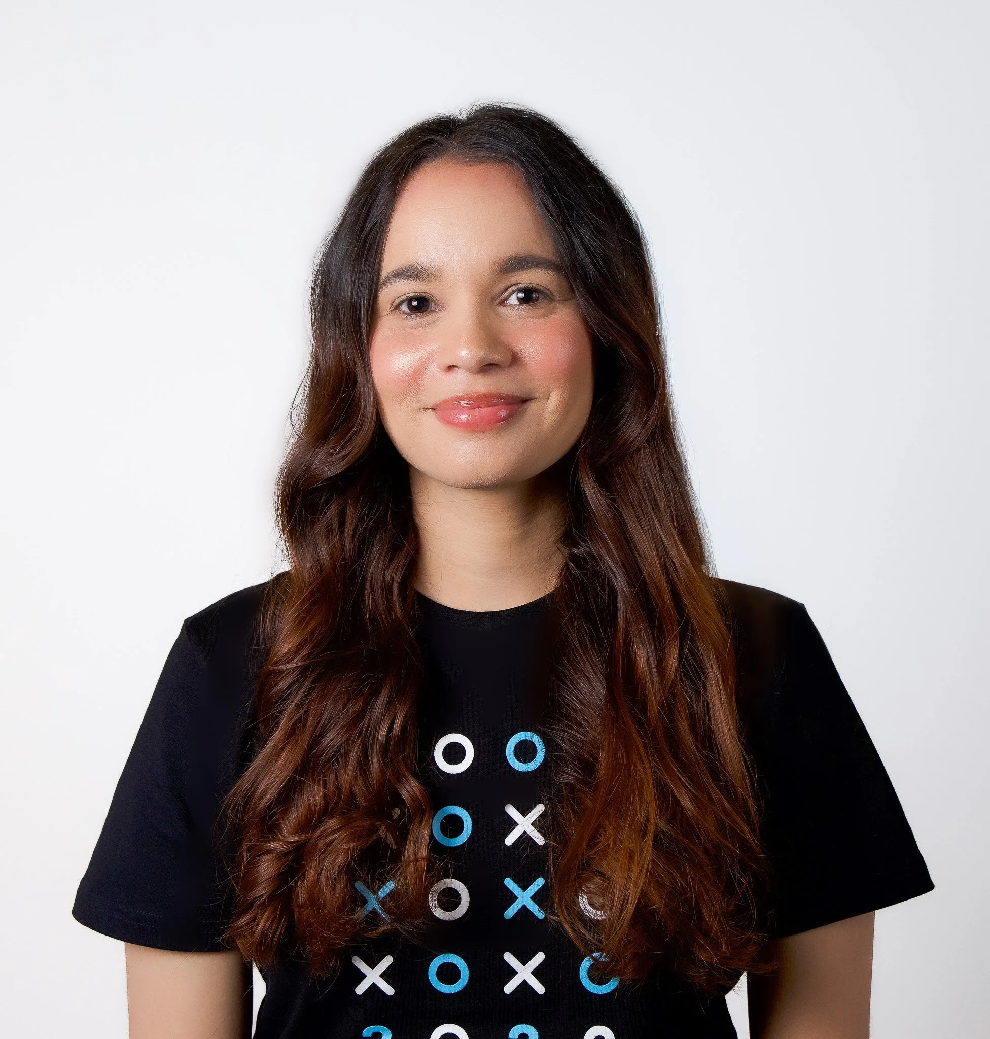 Close-up of a woman with long wavy brown hair, smiling, wearing a black t-shirt with white and blue circles and crosses, against a plain white background.