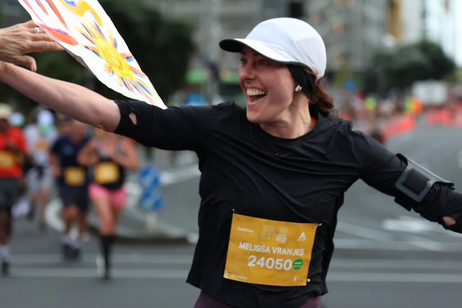 A woman with a white cap and black athletic wear smiling and reaching out to high-five someone during a race, with a bib number 24050 and name Melissa Vranjes, while other runners are visible in the background.