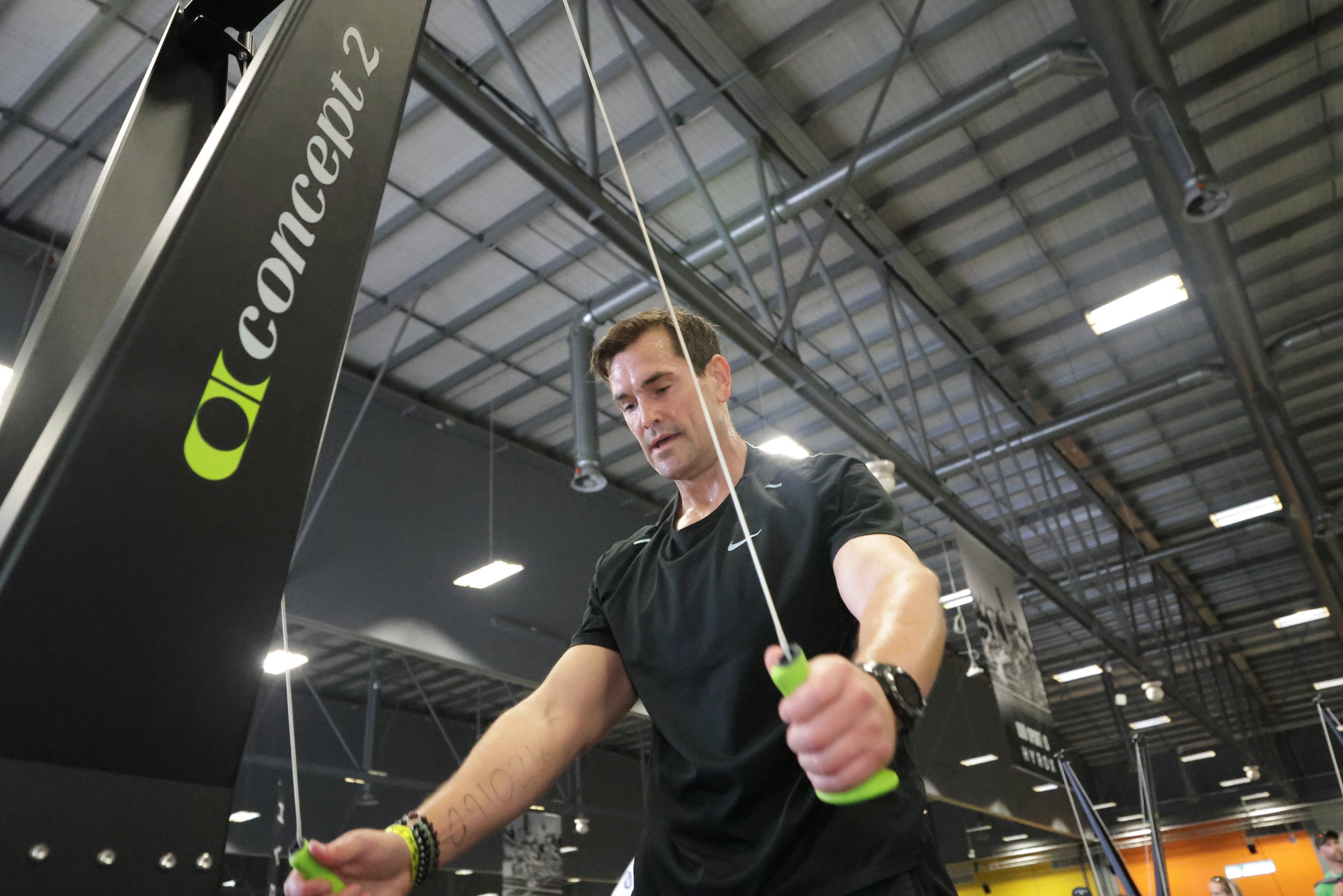 A man working out at a gym, wearing a black T-shirt and a watch, using resistance bands with a gym logo in the background.