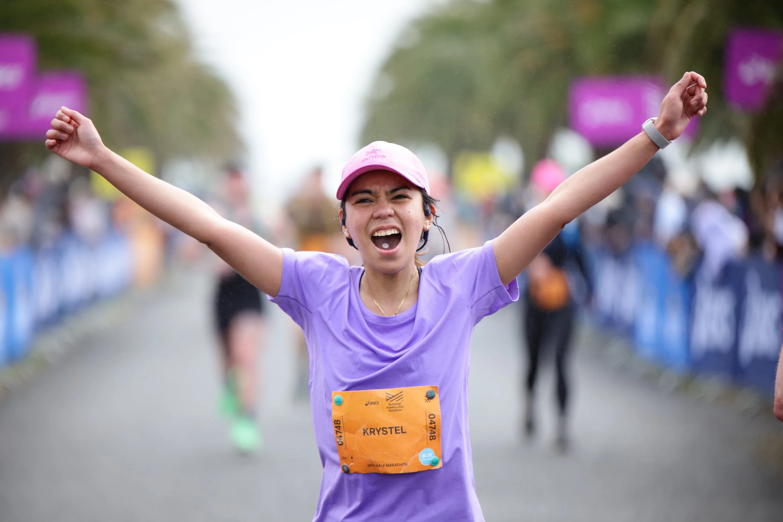 A young woman wearing a pink cap and purple shirt, with a bib that says 'Krystel,' celebrating as she passes the finish line in a race, with her arms raised in victory, surrounded by other runners in a street setting.