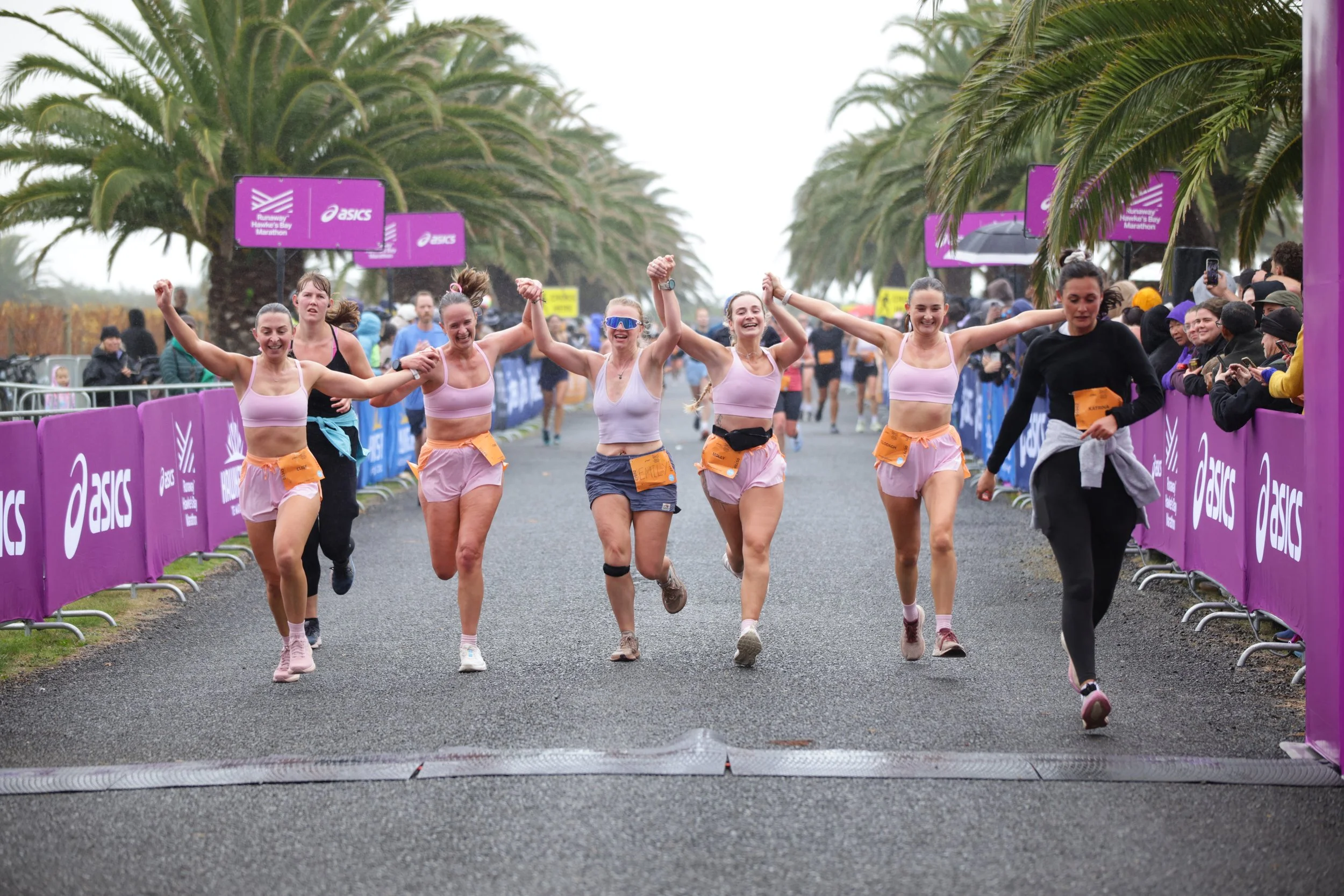 Women crossing finish line at a marathon, smiling, holding hands with arms raised in victory, with spectators and palm trees in the background.