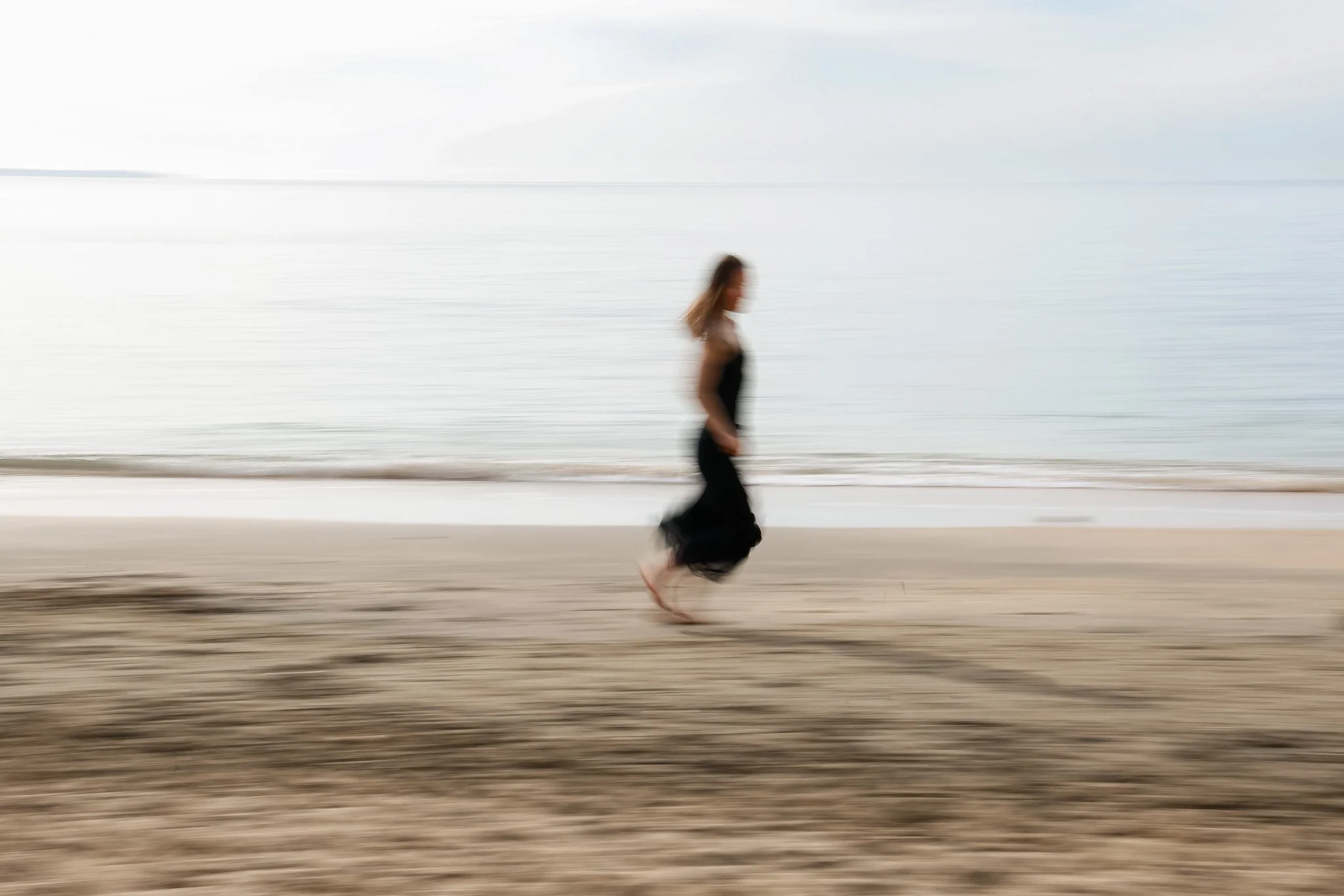 A woman walking barefoot on a beach with blurred background of water and sky.