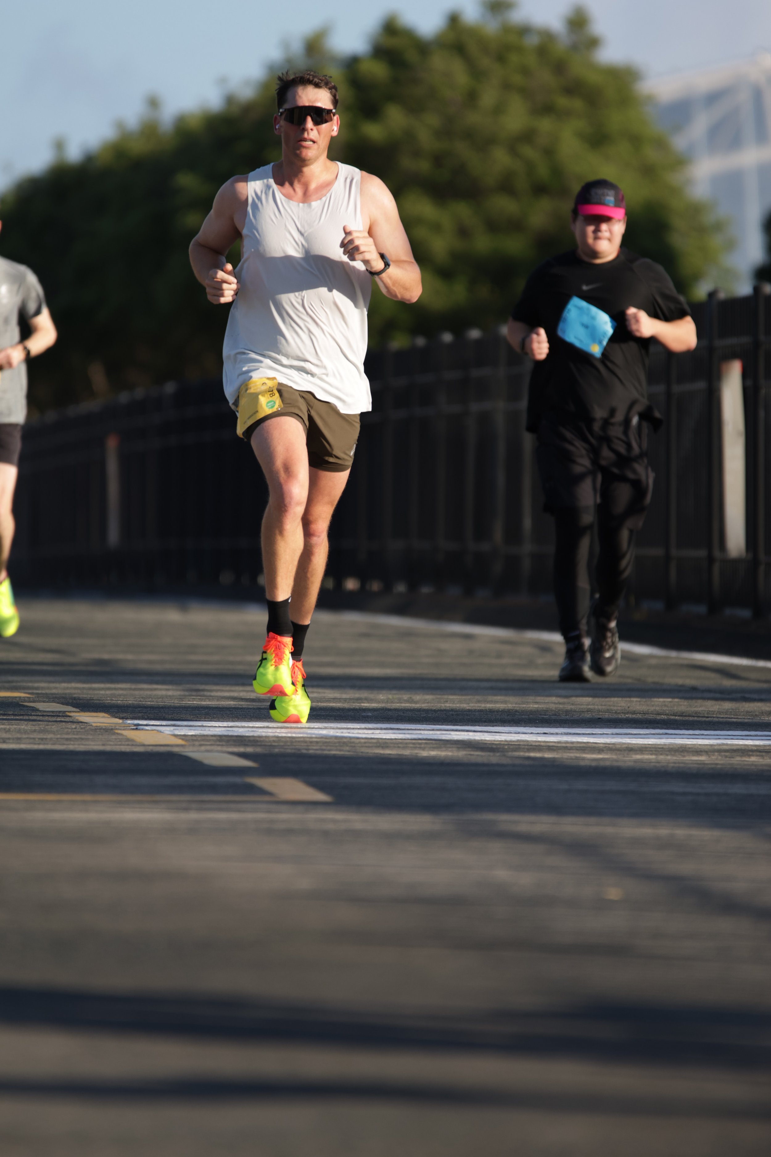 Two runners participating in a race, running on a road with a black fence on the side, with trees and a bridge in the background, in bright daylight.
