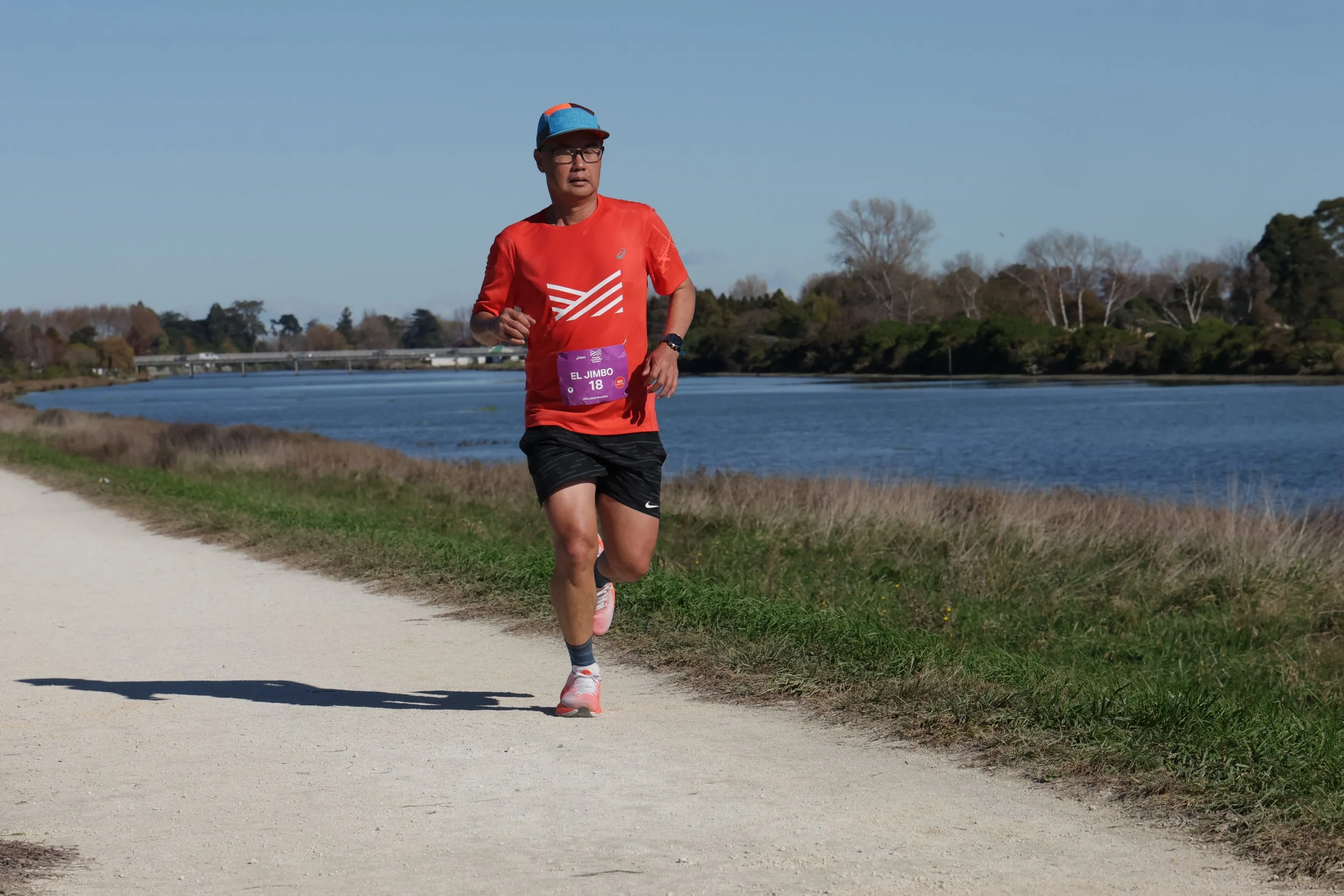 A man running outdoors near a river, wearing an orange shirt, black shorts, a blue cap, glasses, and a marathon bib numbered 18.