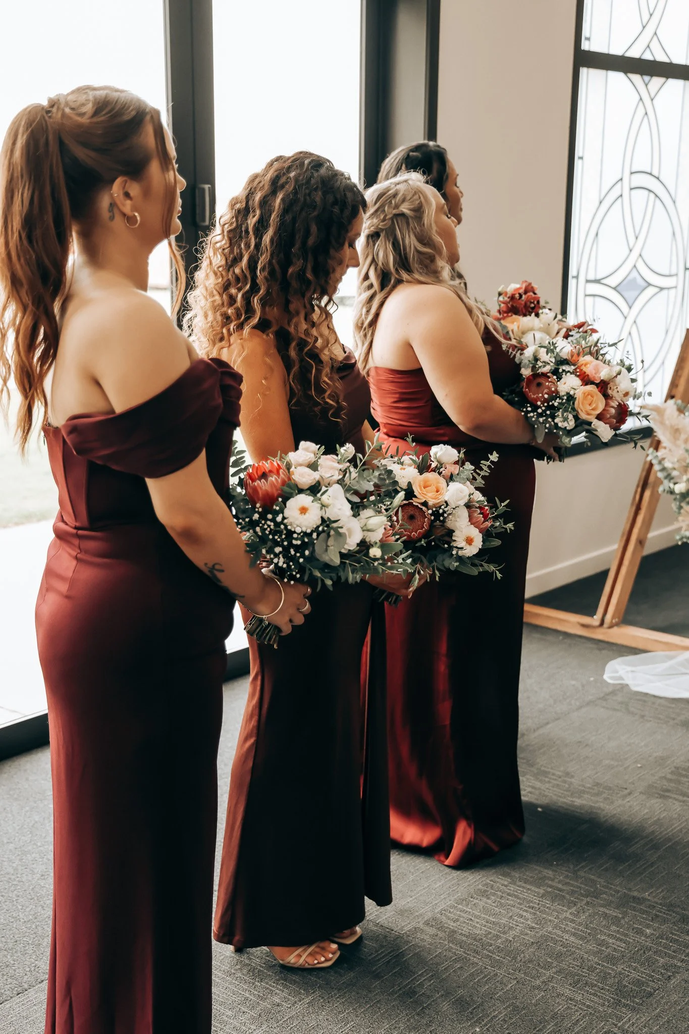 Four women dressed in burgundy gowns holding bouquets of flowers, standing in a line during a wedding ceremony.