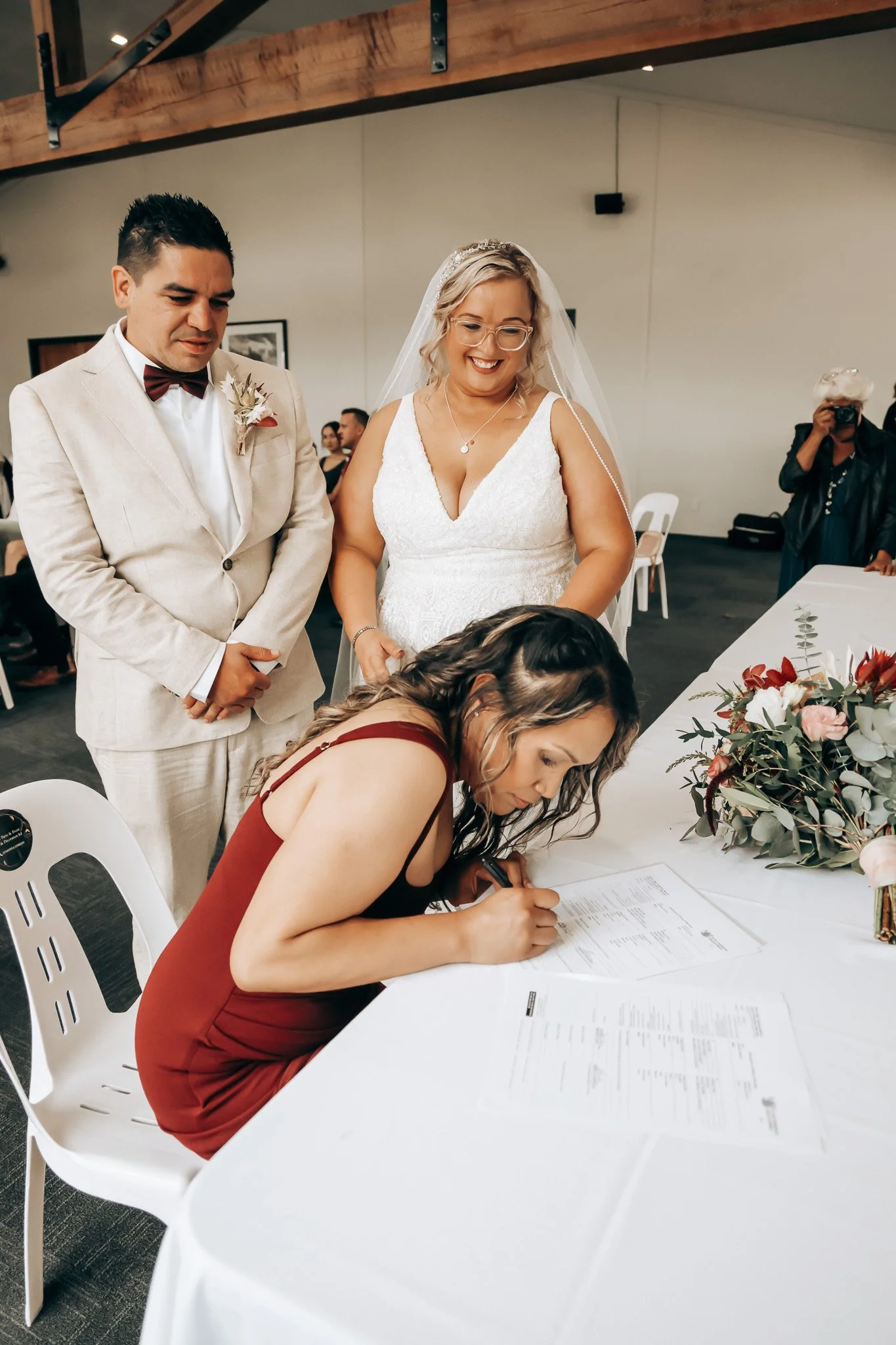 A woman in a red dress signing a document at a wedding reception, with a bride and groom standing nearby, and other guests in the background.