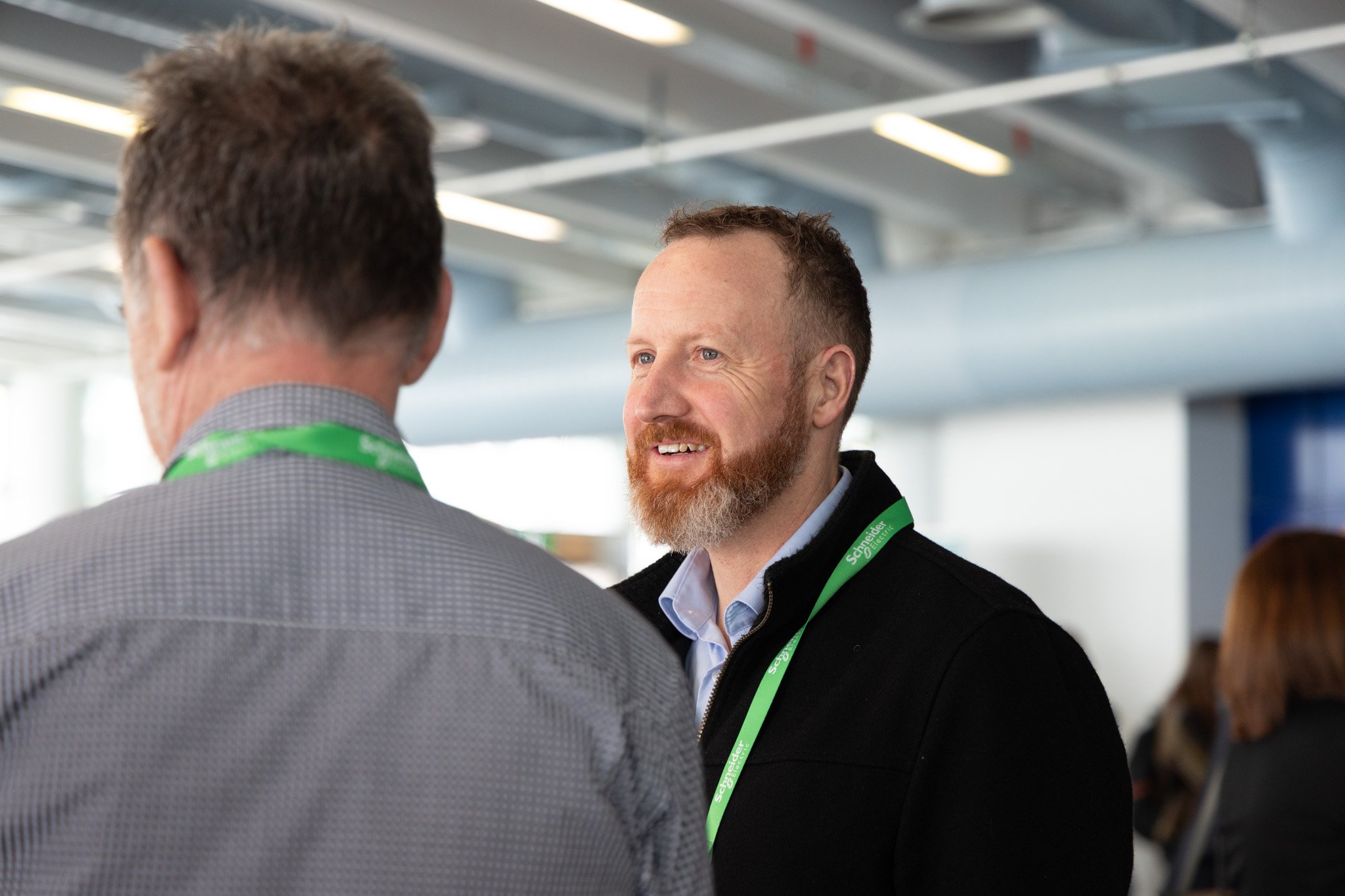 Two men having a conversation in an indoor setting. One is shown from behind, wearing a gray checkered suit and a green lanyard, while the other has a beard, short brown hair, and is dressed in a black jacket with a light blue shirt and a green lanya