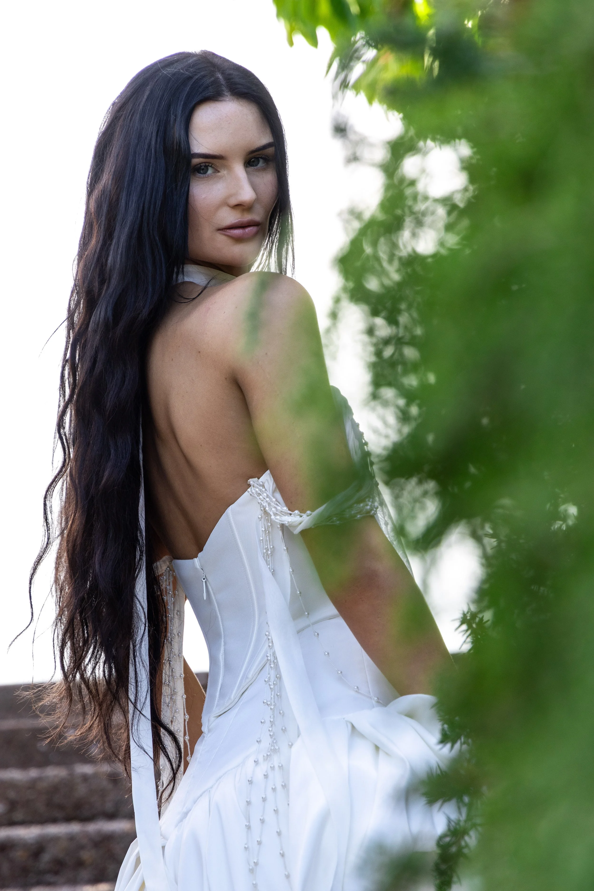 A woman with long dark hair and fair skin, wearing a white dress with intricate beading, standing outdoors near green foliage and stairs, looking at the camera.