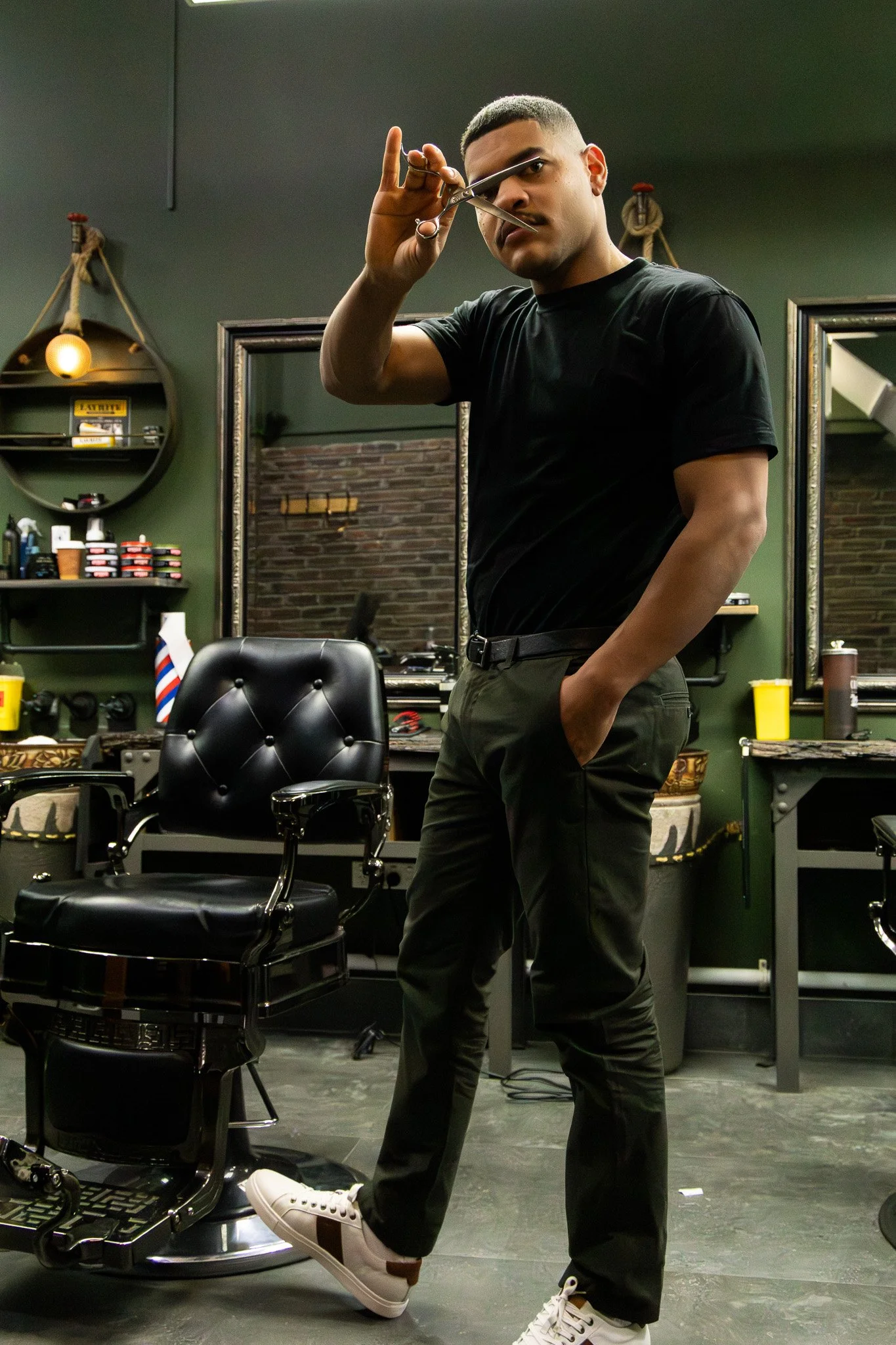 A man with short hair and a mustache wearing a black T-shirt and black pants, holding a pair of scissors and posing in a barbershop or salon. The background features mirrors, barber tools, and shelves with products, with a dark brick wall visible beh