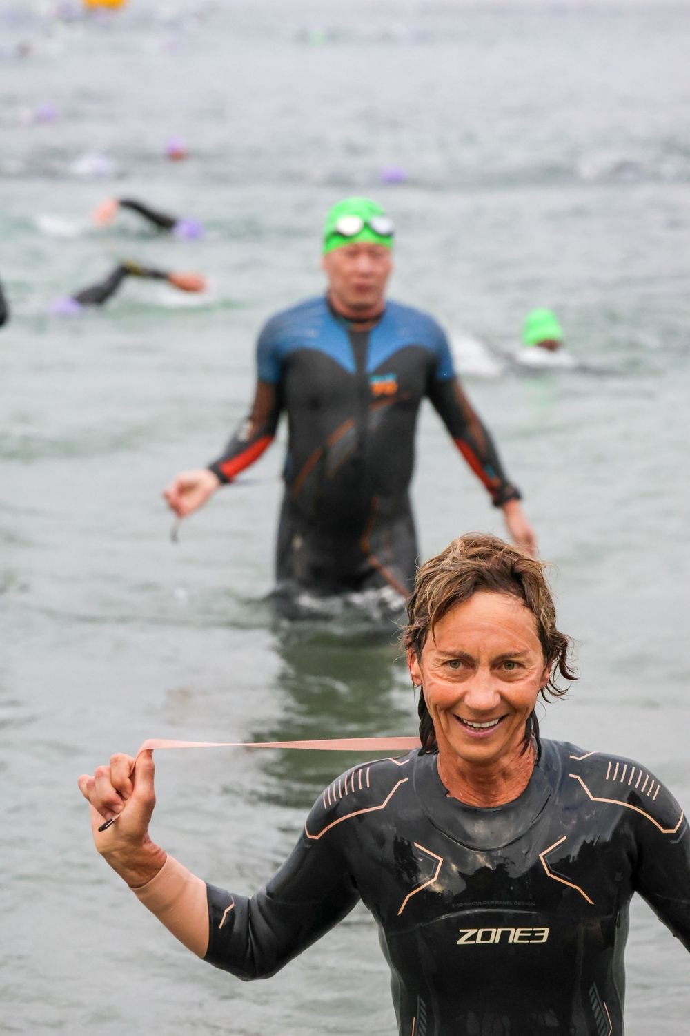 A group of people participating in a triathlon swimming in open water. The person in the foreground is smiling and holding a pink strap, with others swimming behind him.