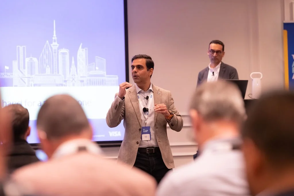 Men giving a presentation at a conference with a large screen displaying a city skyline in the background.
