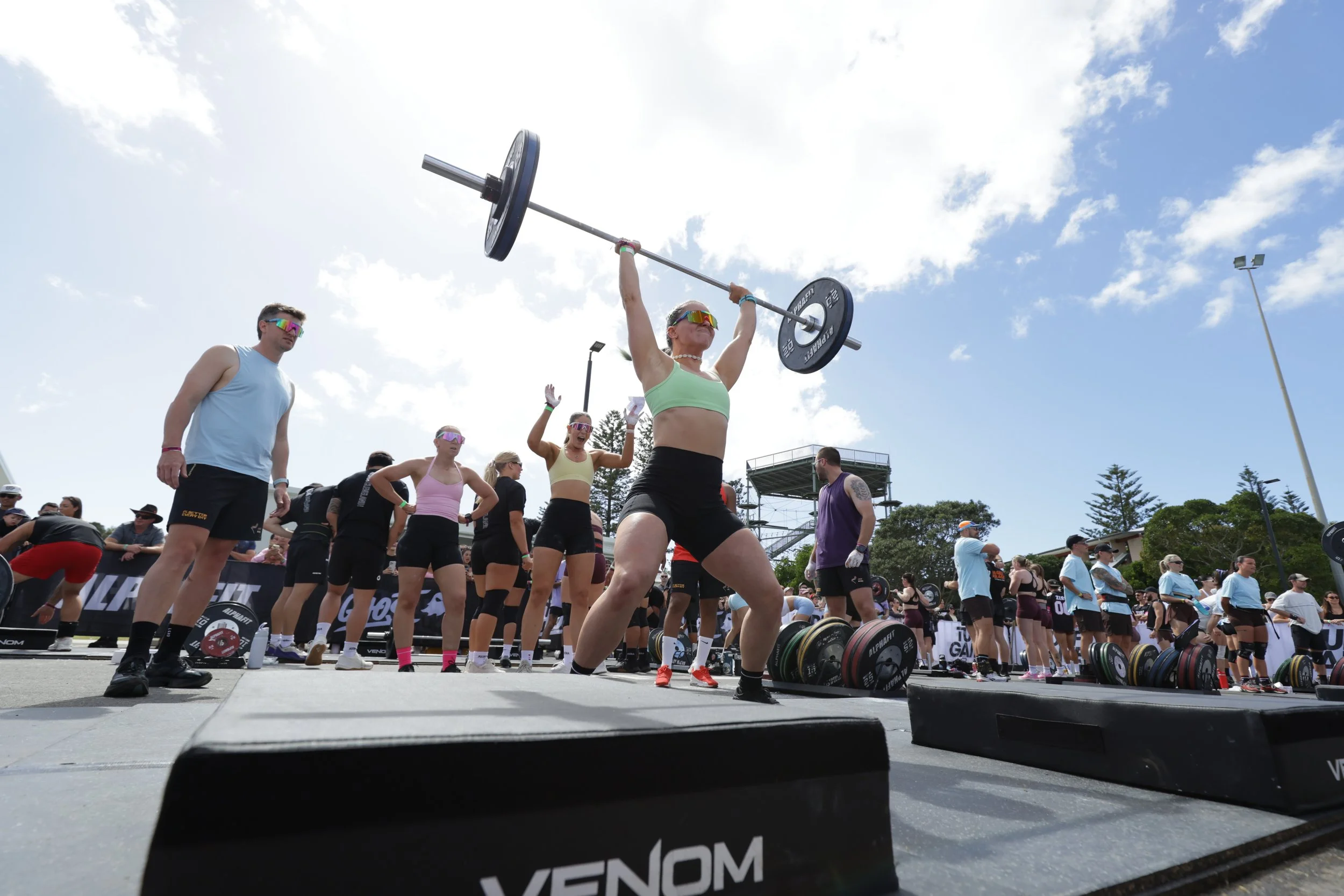 A woman in a green sports bra and black shorts lifts a barbell overhead at a CrossFit outdoor competition, surrounded by other athletes and spectators under a partly cloudy sky.