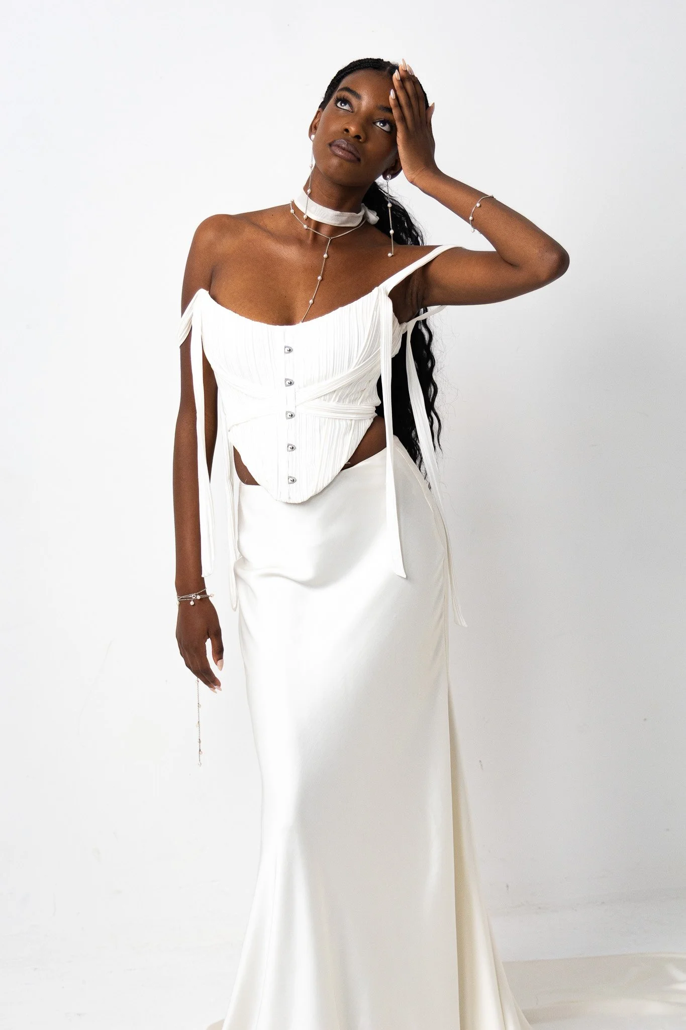 A woman with dark skin and long wavy black hair posed in an elegant white dress against a plain white background. She is accessorized with layered necklaces, bracelets, and earrings, and has her right hand on her forehead with her eyes looking upward