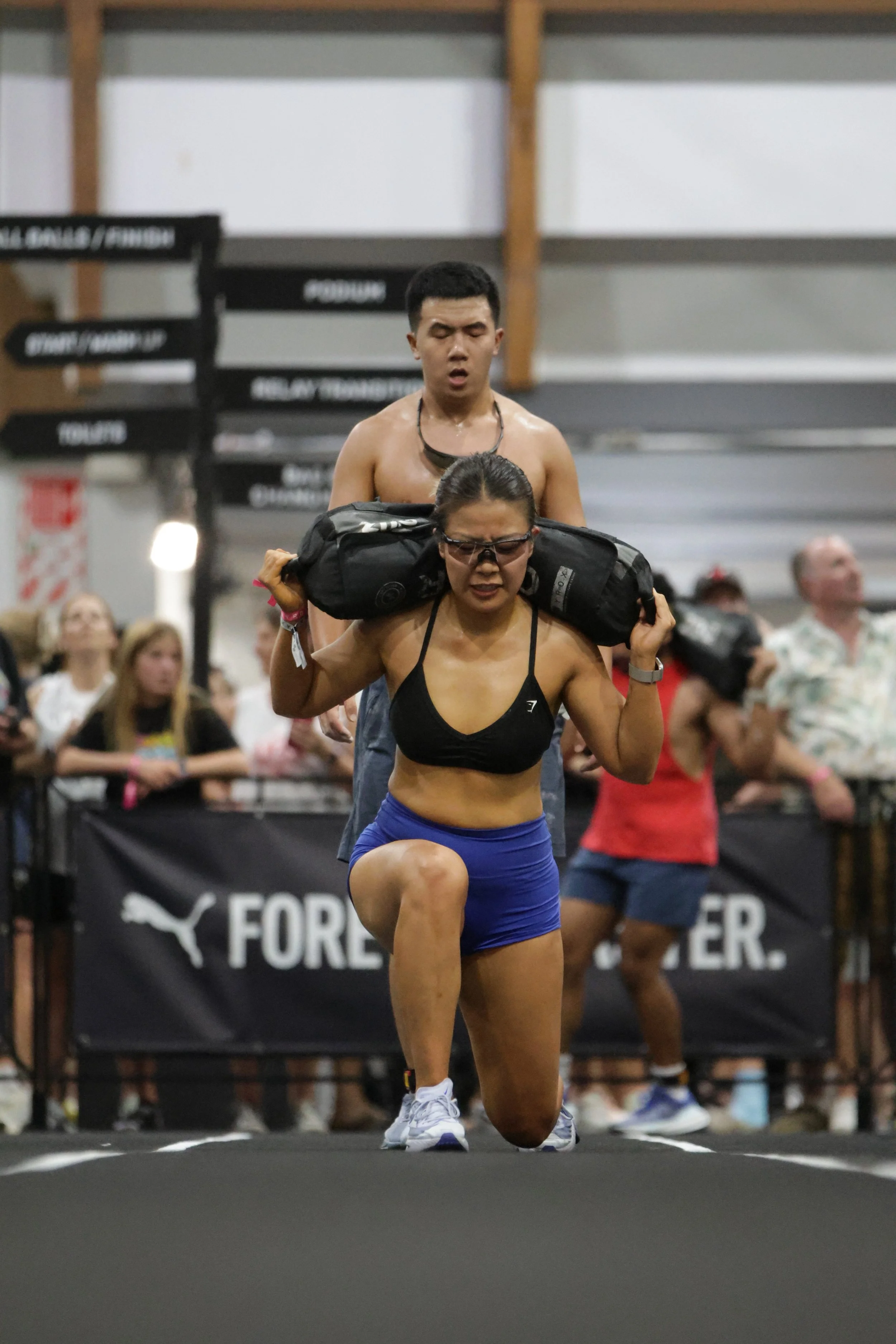 A woman in athletic gear performs a lunge exercise while a man with a black bag over his shoulders stands behind her during a fitness event, with spectators in the background.