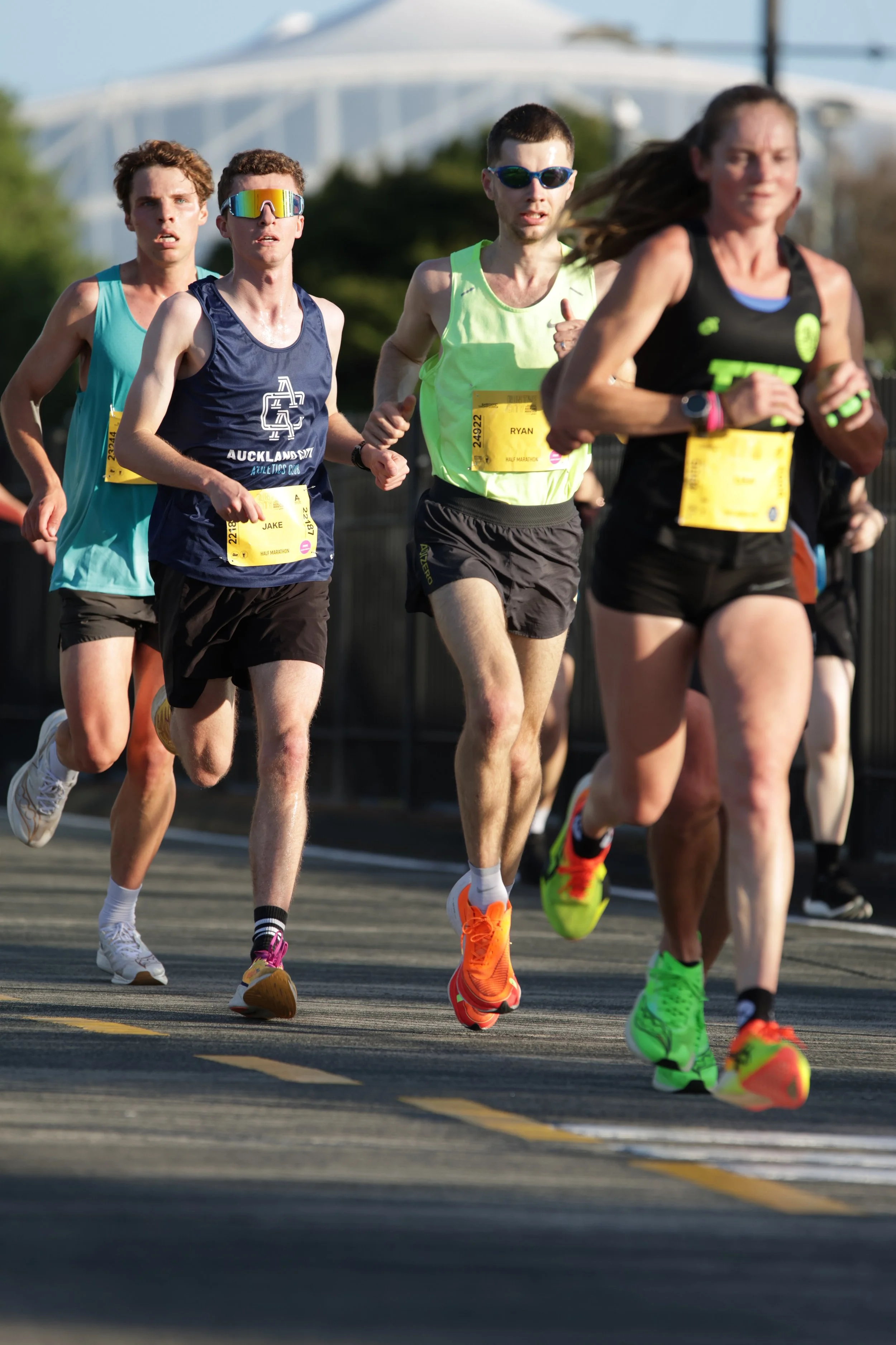 Group of runners participating in a race, wearing athletic gear and sunglasses, running on a paved road with a bridge in the background.