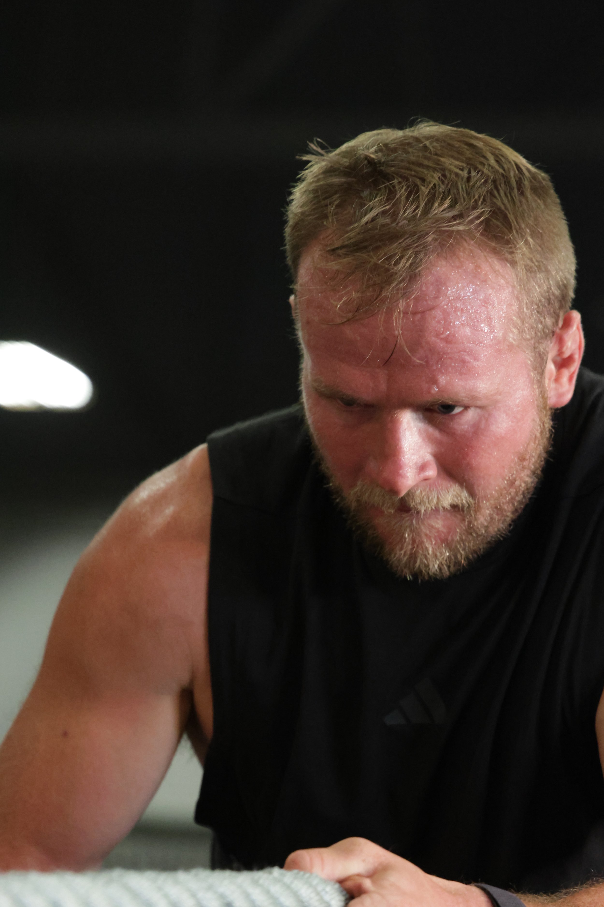 A man with blond hair and a beard, wearing a black sleeveless shirt, appears to be working out intensely in a gym.