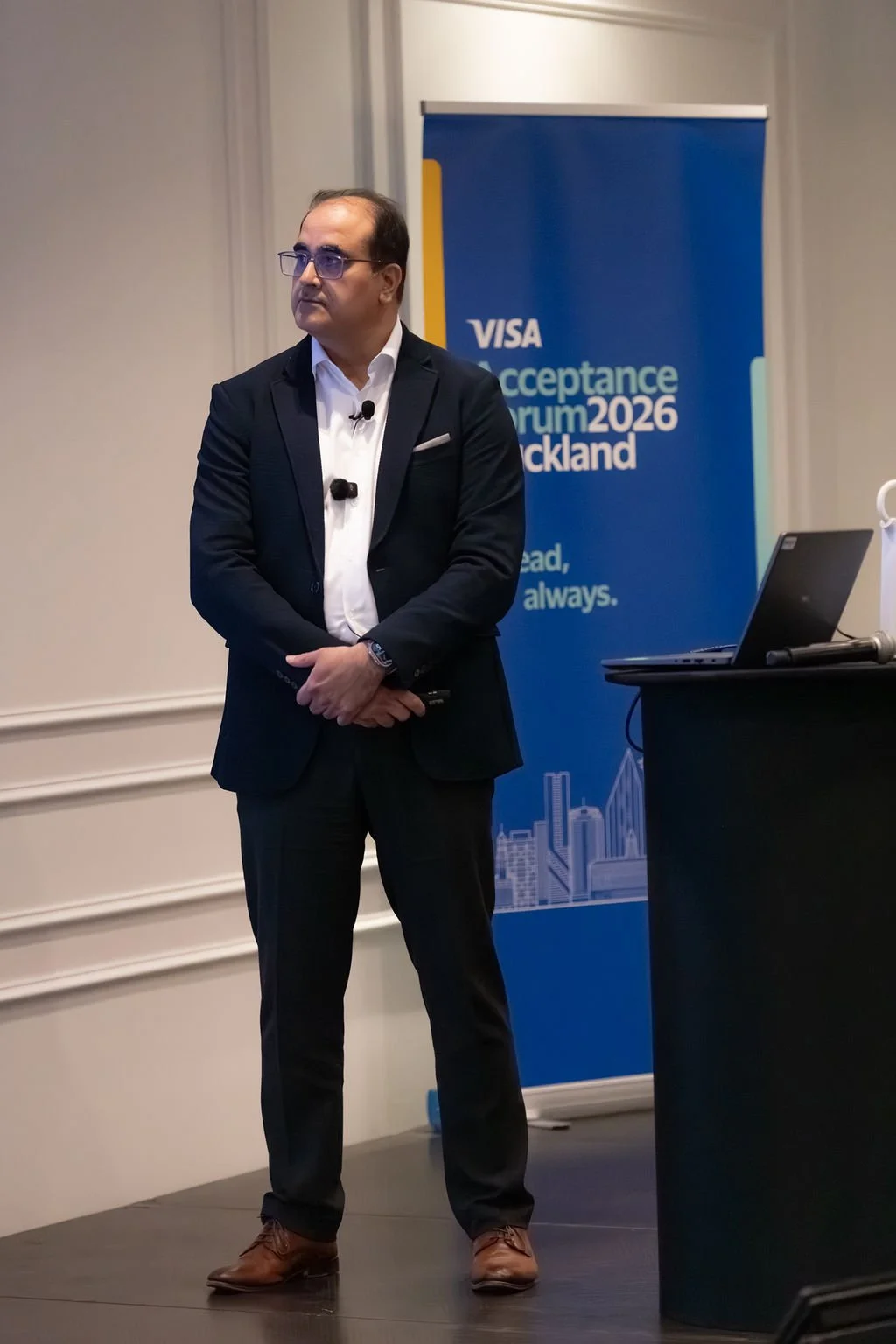 A man in a dark suit and glasses standing on stage near a blue banner that reads 'Visa Acceptance Forum 2026 Auckland'. He is holding a microphone and has a laptop on a black table beside him.