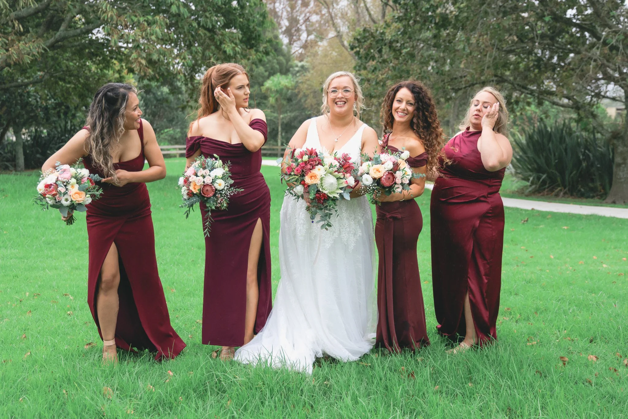 Group of five women standing together outdoors on a grassy area, dressed in formal attire, holding bouquets of flowers, with one in a white wedding dress, smiling and laughing.