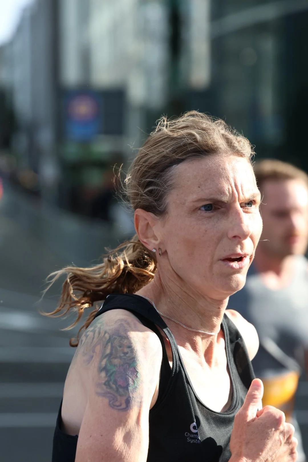 A woman with light skin and tattoos running on a city street in daylight, wearing a black athletic top.