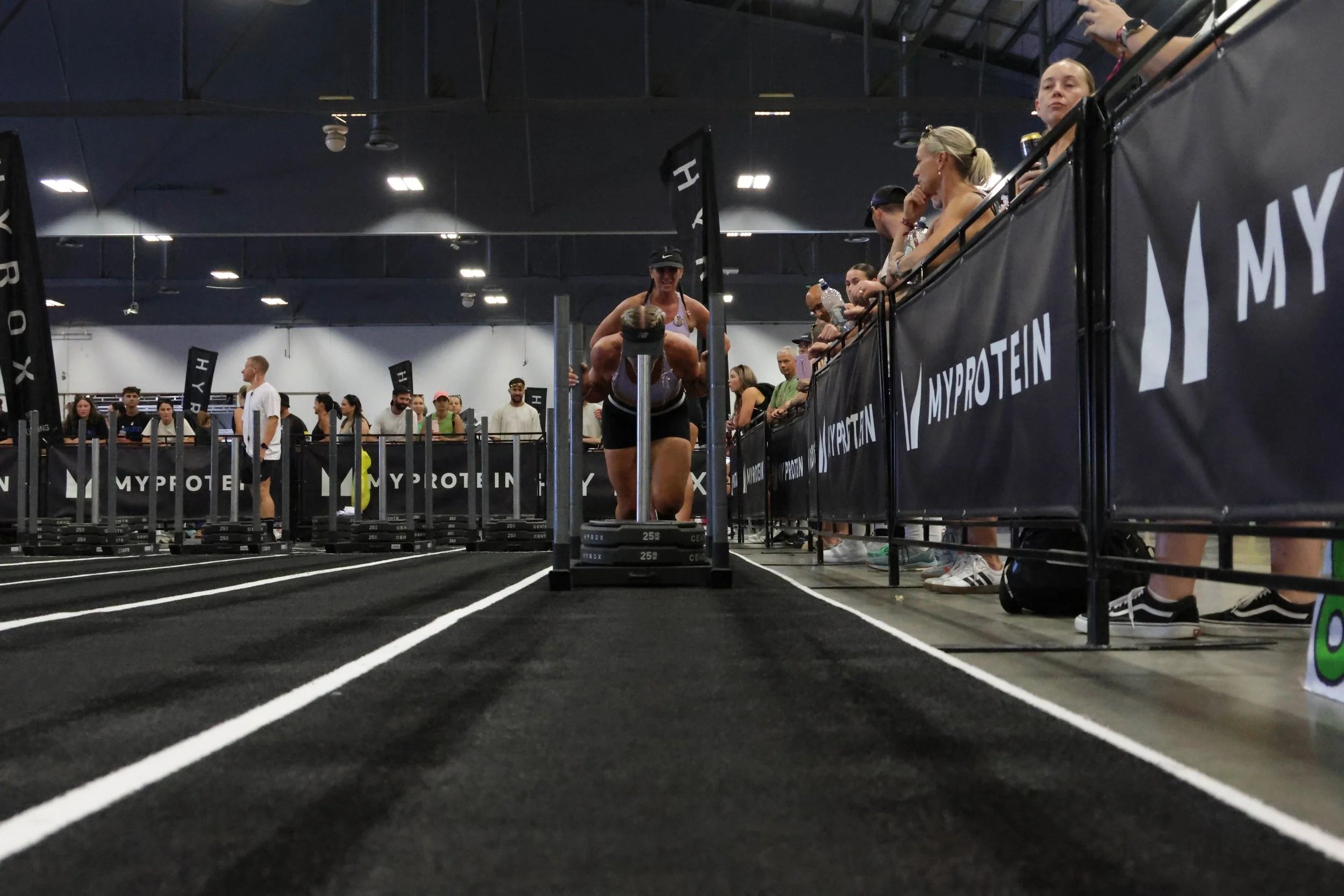 A group of women waiting behind a black barrier at an indoor athletic event, with some adjusting their hair and others observing the activity on the track, which is set up for a race or exercise.
