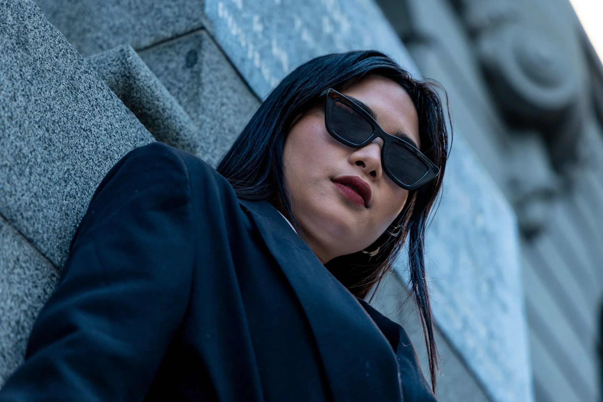 Close-up of a woman with dark hair and sunglasses lying on a stone or concrete surface outdoors.