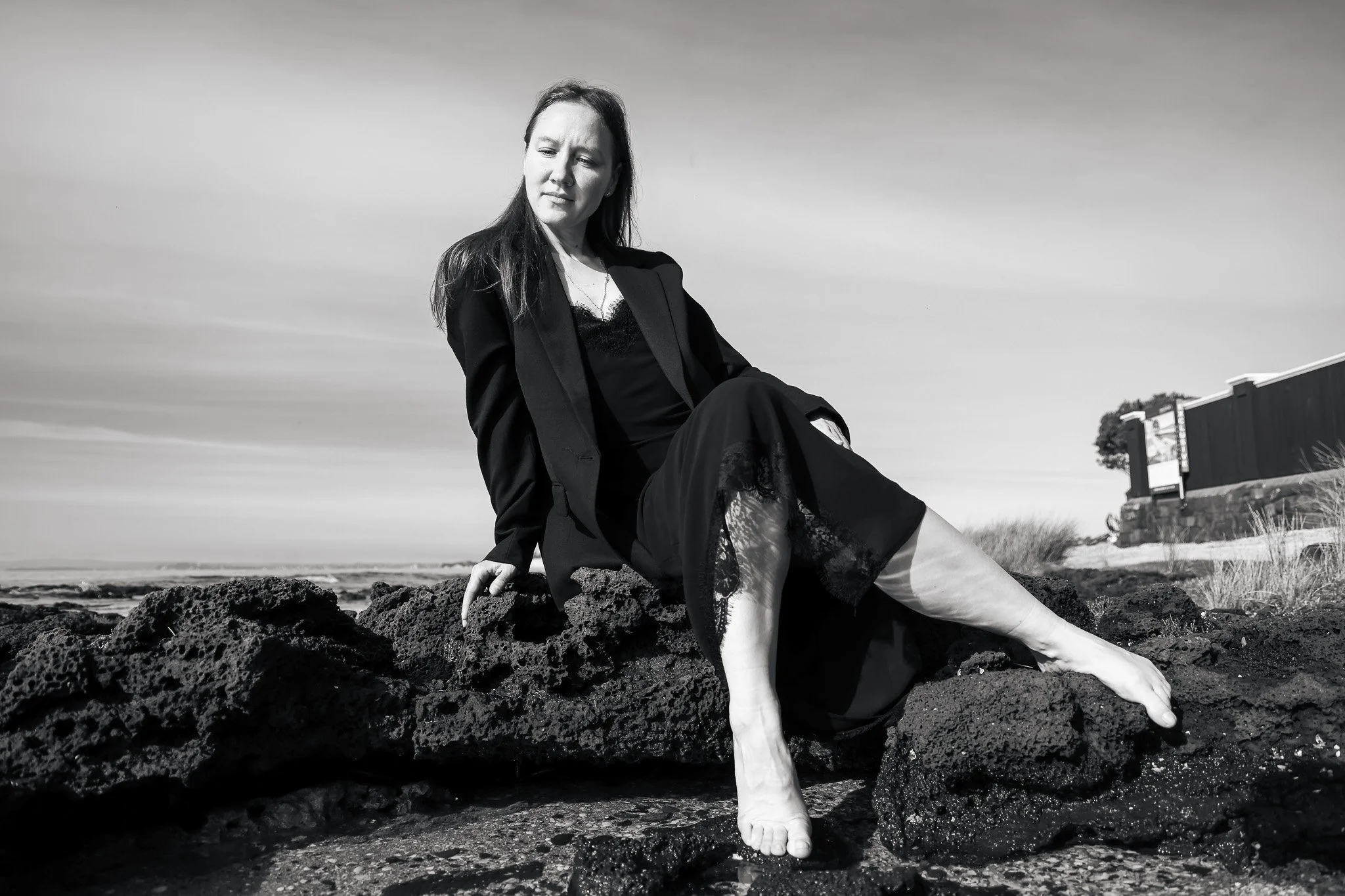 A black-and-white photo of a woman sitting on rocks outdoors, wearing a blazer and lace dress, with a slightly cloudy sky and a building in the background.